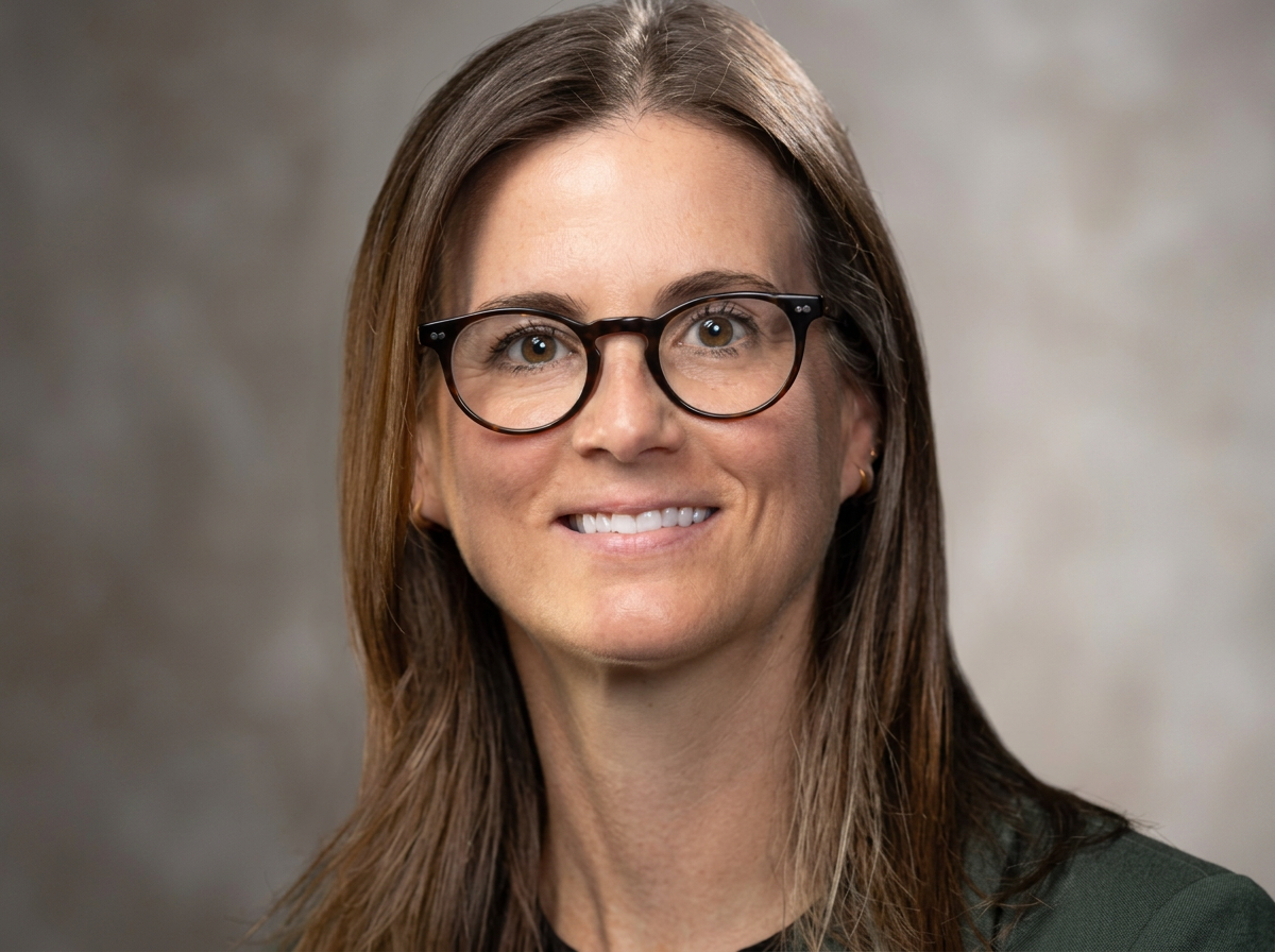 A woman with long brown hair wearing glasses and a dark green top smiles at the camera against a soft gray background.