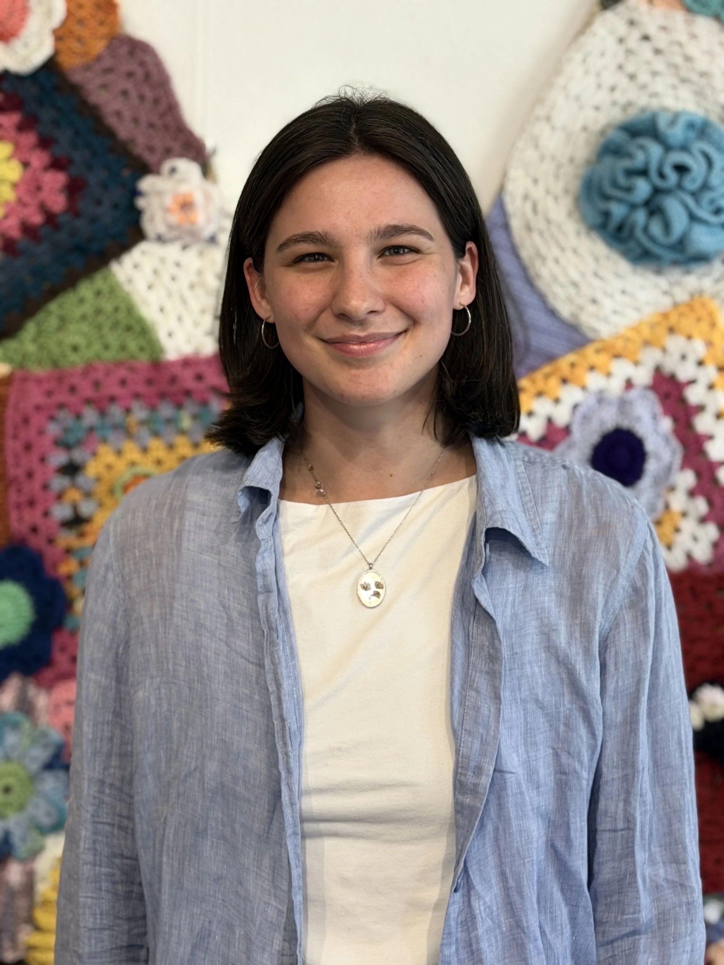 A woman with dark hair smiling, wearing hoop earrings, a chain necklace with a pendant, a white shirt, and a light blue button-up shirt, standing in front of a colorful crocheted or knitted quilt.