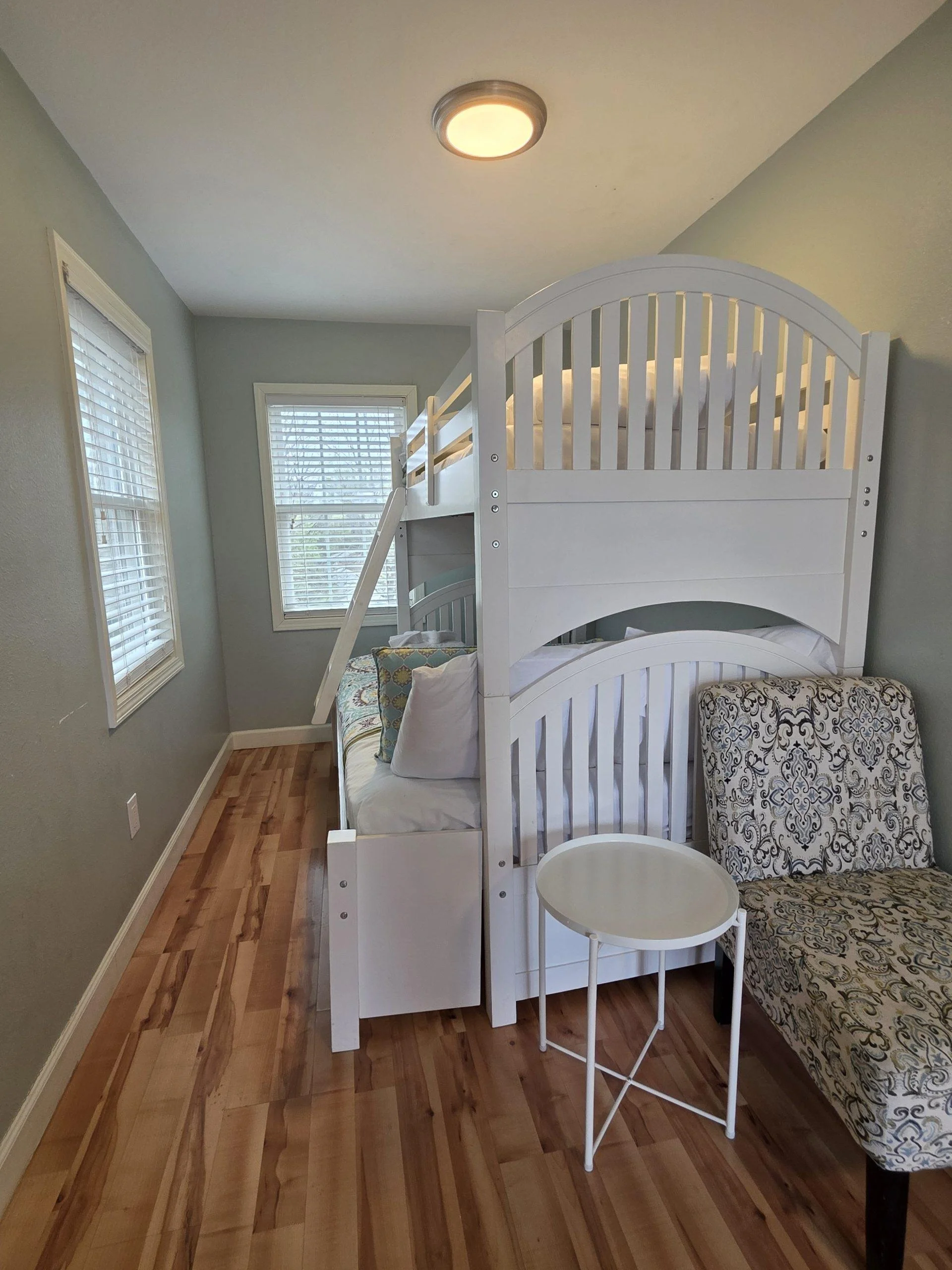 A bedroom corner featuring a white bunk bed, a patterned armchair, a small white side table, and two windows with blinds. The room has light green walls, hardwood floors, and a ceiling light fixture.