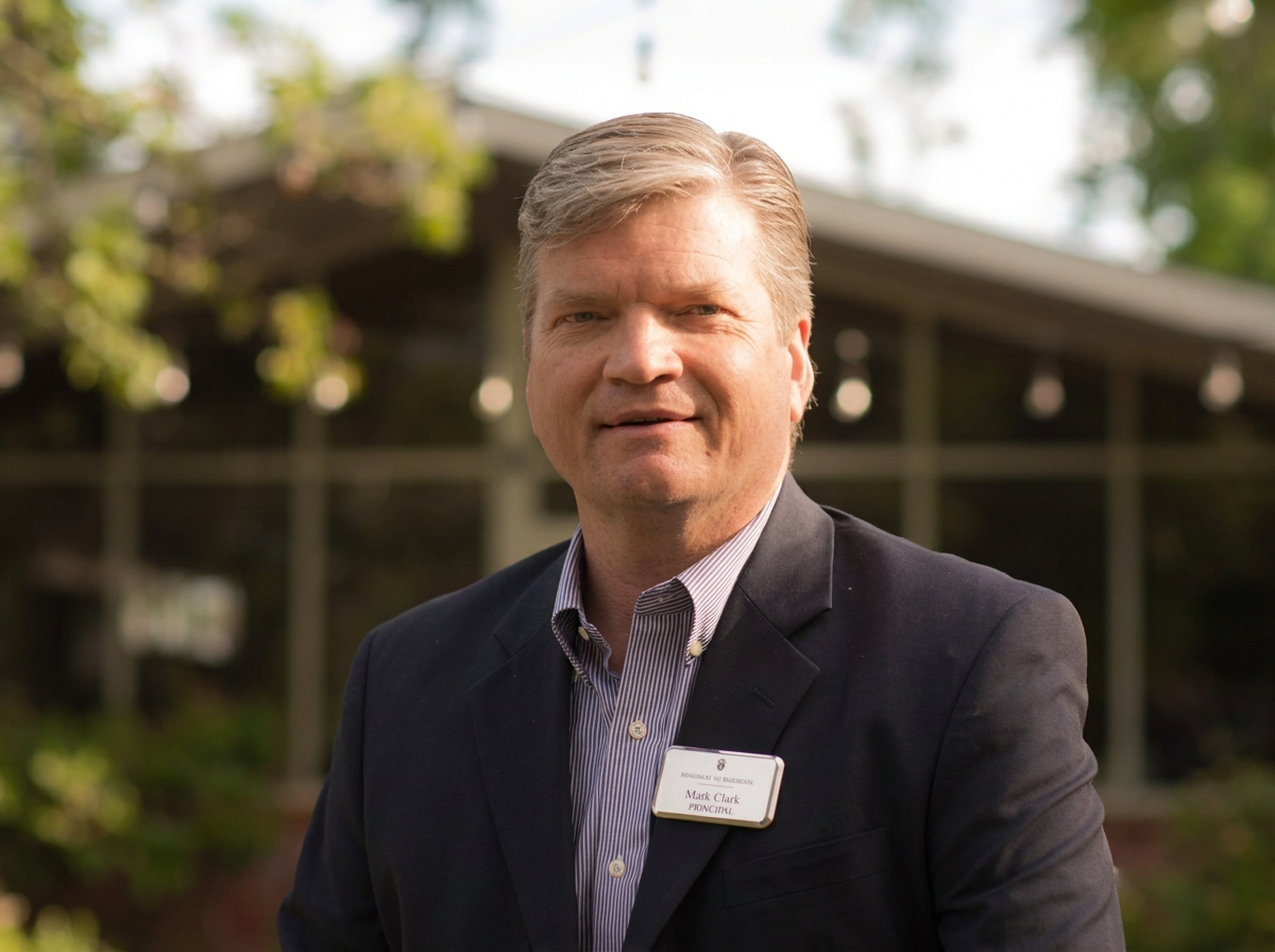 A middle-aged man with blonde hair wearing a dark blazer and a striped dress shirt, standing outdoors with trees and a house in the background.