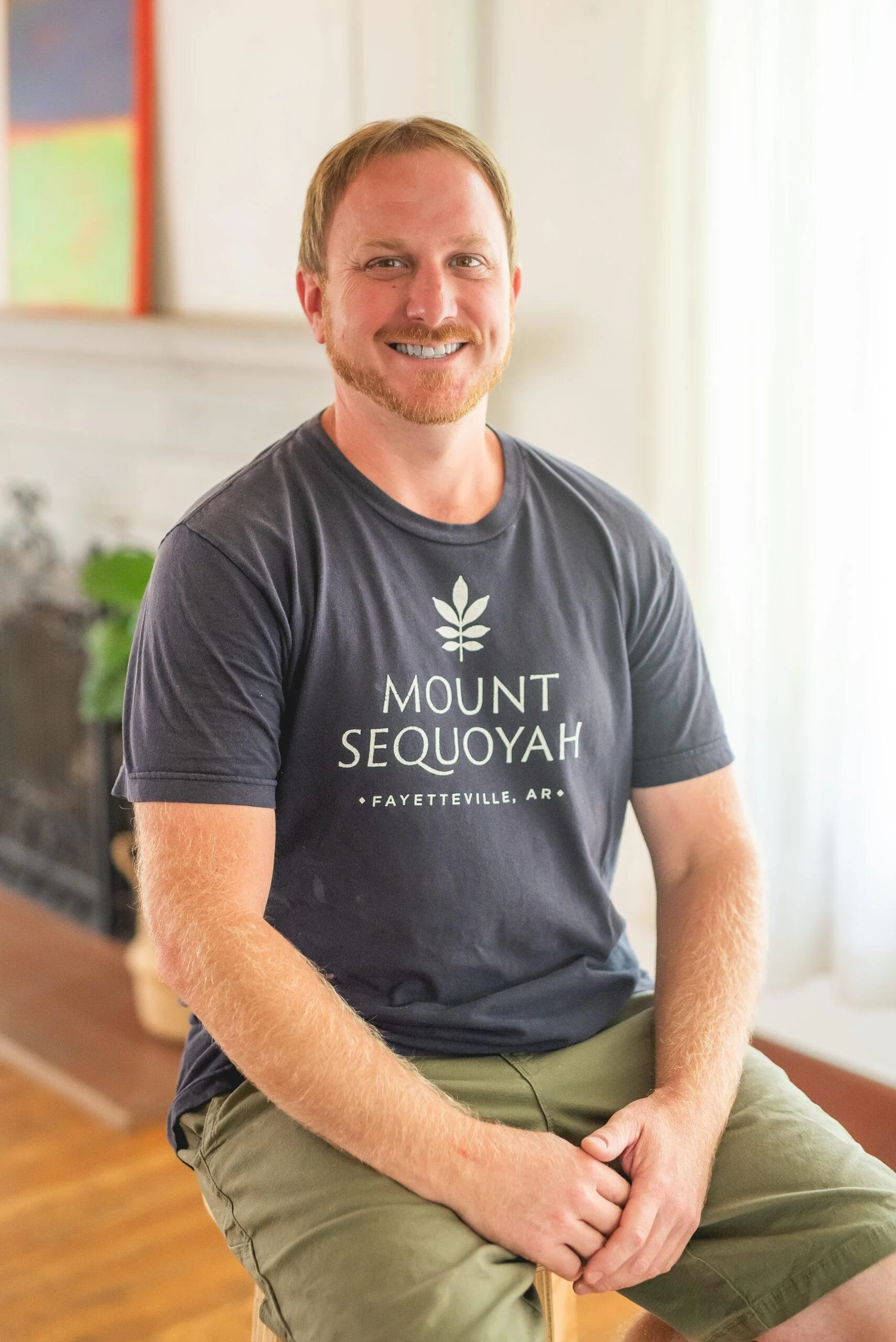A smiling man with a red beard and mustache, wearing a dark t-shirt with the words 'Mount Sequoyah Fayetteville AR' and sitting indoors with a window behind him.