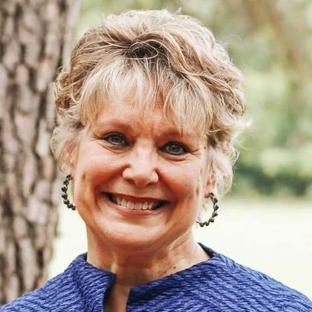 Close-up of a smiling middle-aged woman with short blonde hair, wearing hoop earrings and a blue textured jacket, outdoors near a tree.