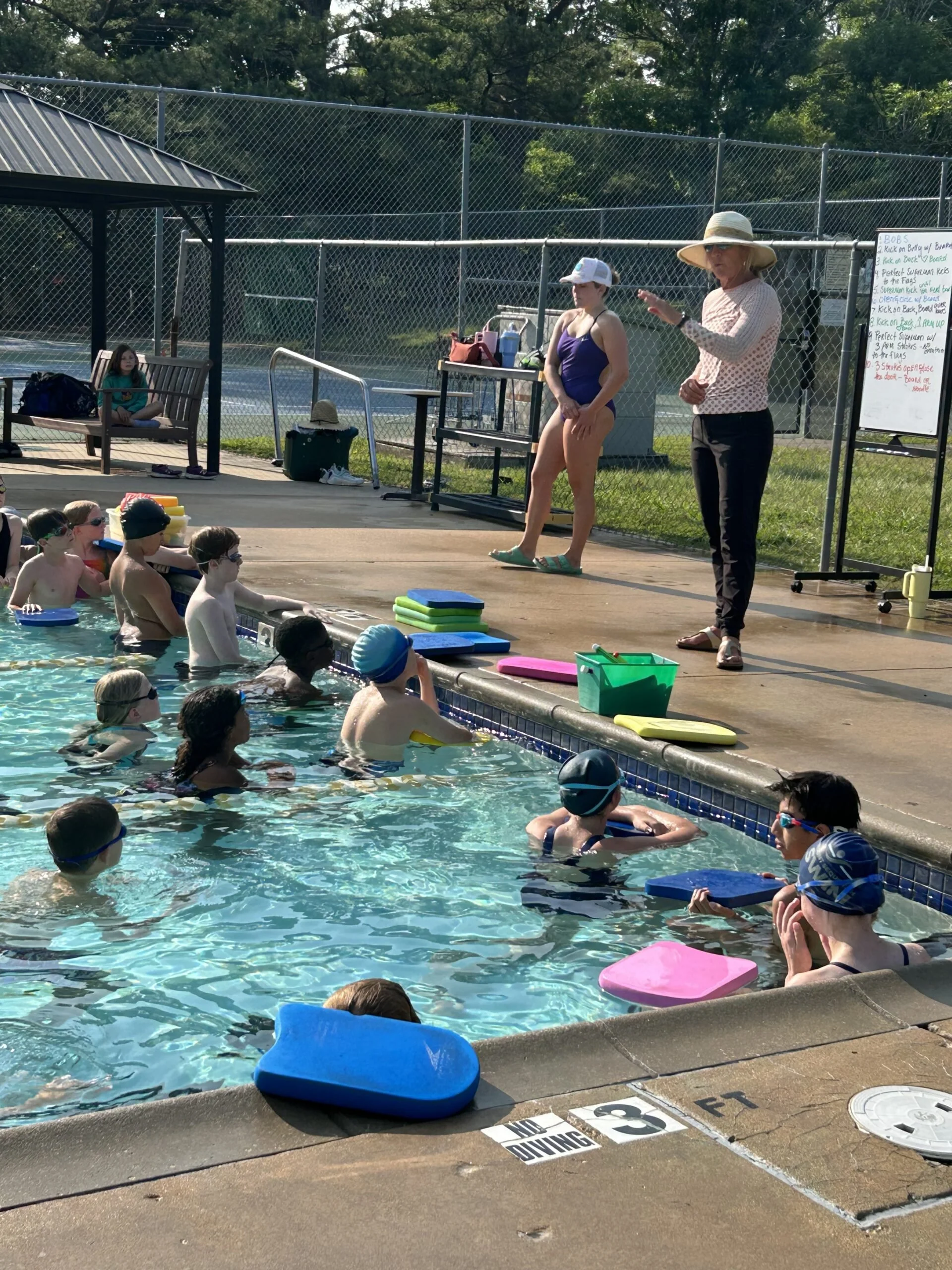 Children participating in a swimming class at an outdoor pool, with an instructor coaching them. Some kids are in the water with floating boards, while others are sitting on the poolside. The pool area is fenced, and there are benches and a whiteboard with instructions visible.