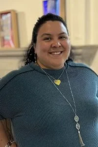A smiling woman with dark hair, wearing a blue top and pendant necklace, standing indoors in front of a bookshelf.