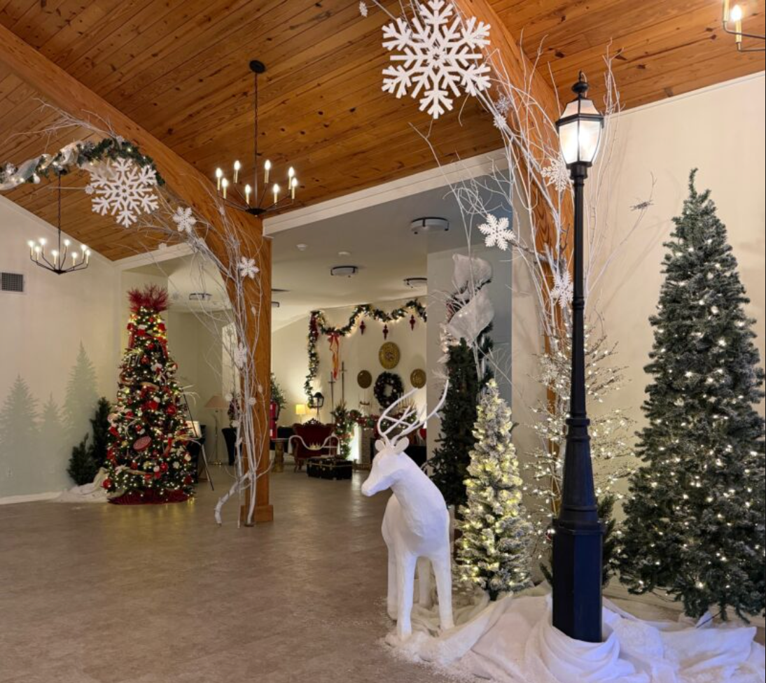 Festive indoor Christmas scene with decorated Christmas trees, a white reindeer figure, snowflake ornaments, and string lights. There are garlands, a lamp post, and holiday decorations on the wall.