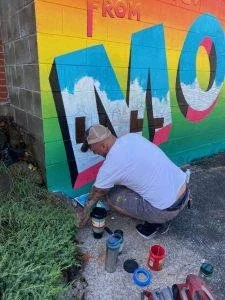 Person spray painting a colorful mural on a wall, with paint cans on the ground nearby.