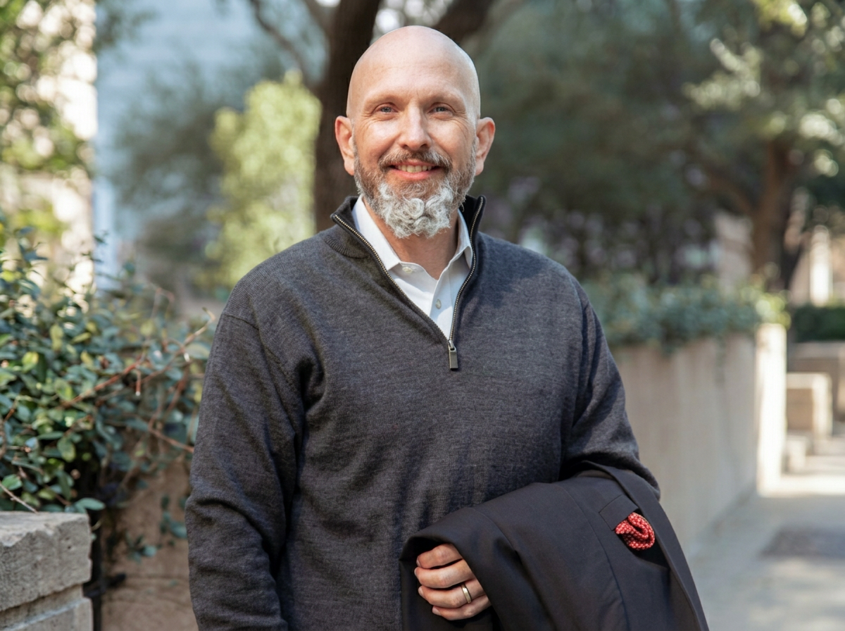 A smiling middle-aged man with a grey beard and bald head, wearing a grey quarter-zip sweater over a white collared shirt, standing outdoors in a park-like setting with trees and a stone wall in the background, holding a black jacket with a red cord in his right hand.
