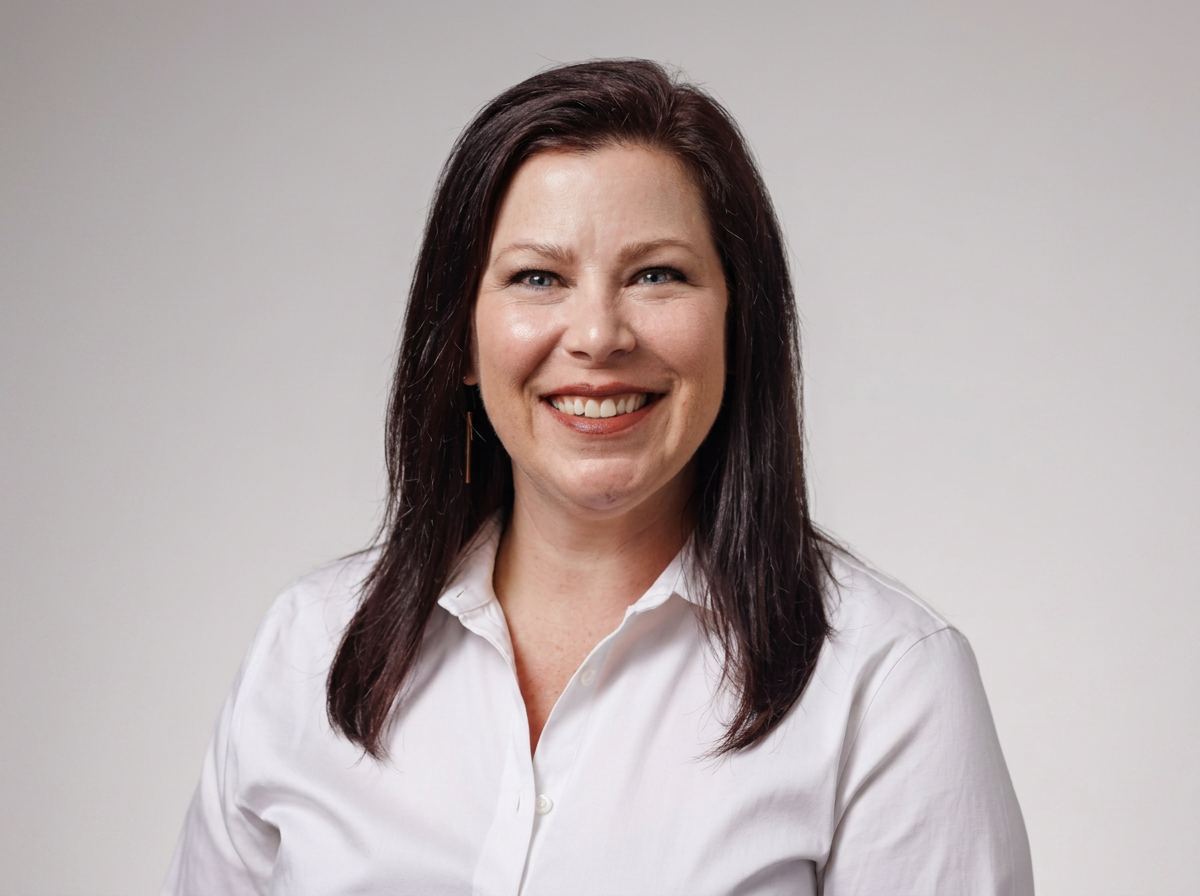 A woman with dark brown hair, wearing a white button-up shirt, smiling against a plain gray background.