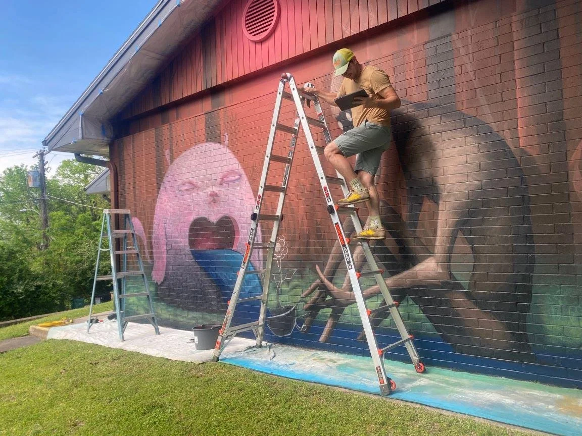 A man standing on a ladder painting a large mural on the side of a brick building, featuring a pink cartoon face with an open mouth and a dog lying on the ground.