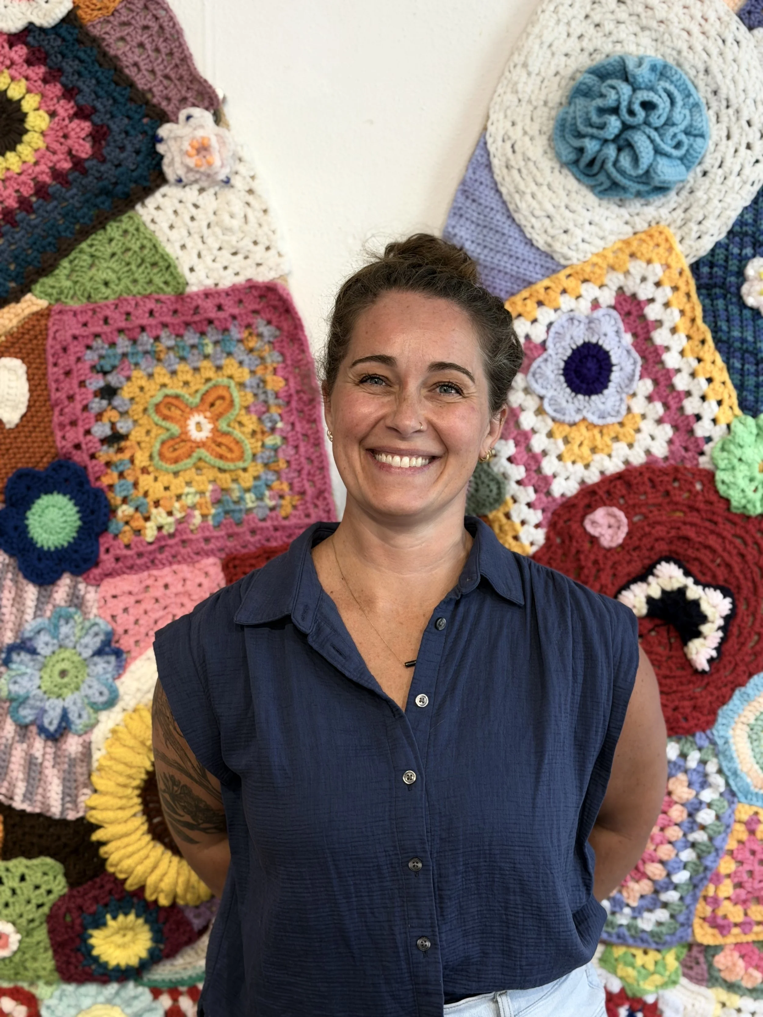 A woman smiling, standing in front of a wall of colorful crocheted and knitted blankets with various patterns and designs.