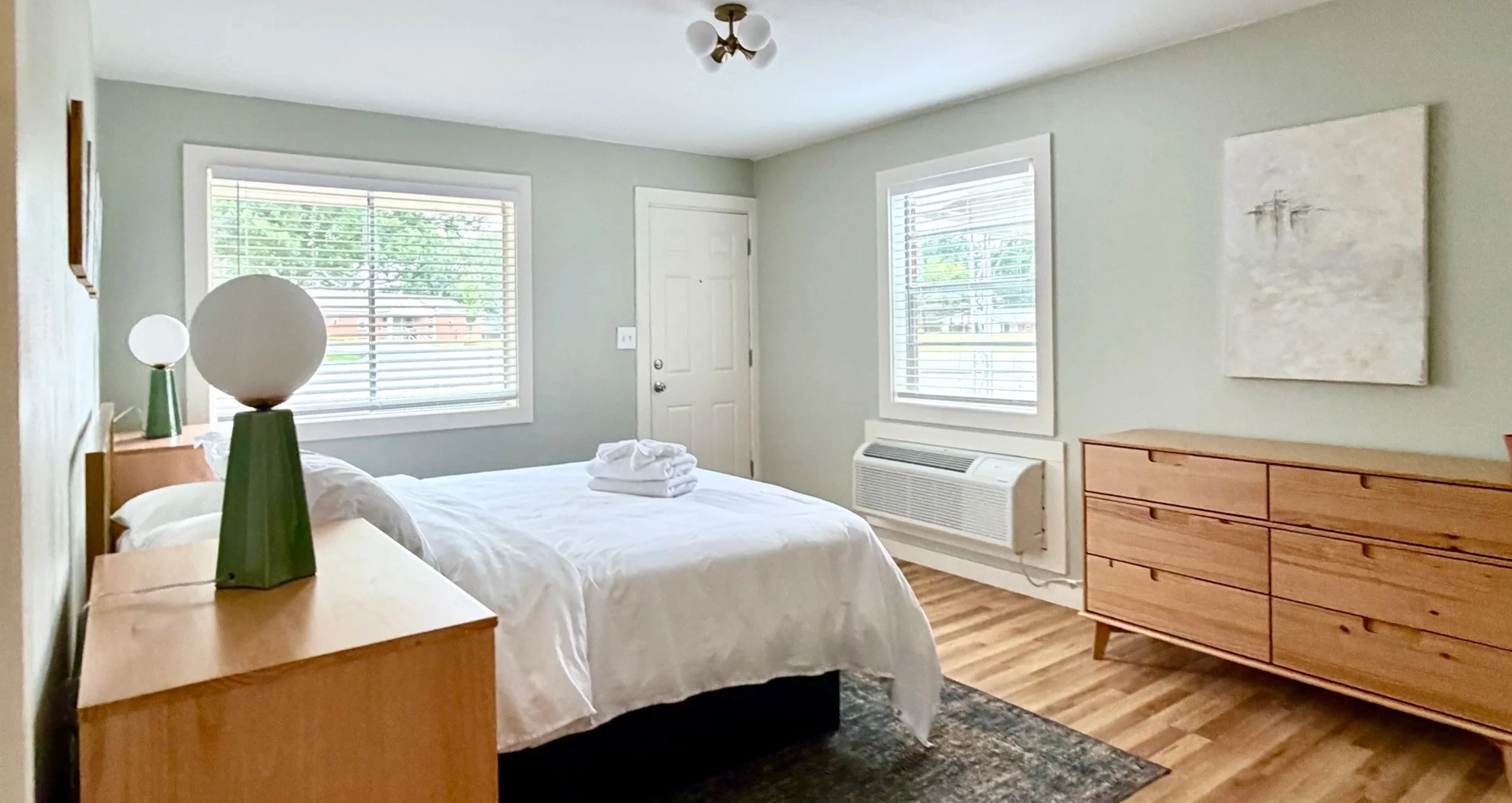 A bedroom with a bed covered in white bedding, a folded towel on top, two windows with blinds, a wooden dresser, a small air conditioning unit below one window, and a modern ceiling light fixture.