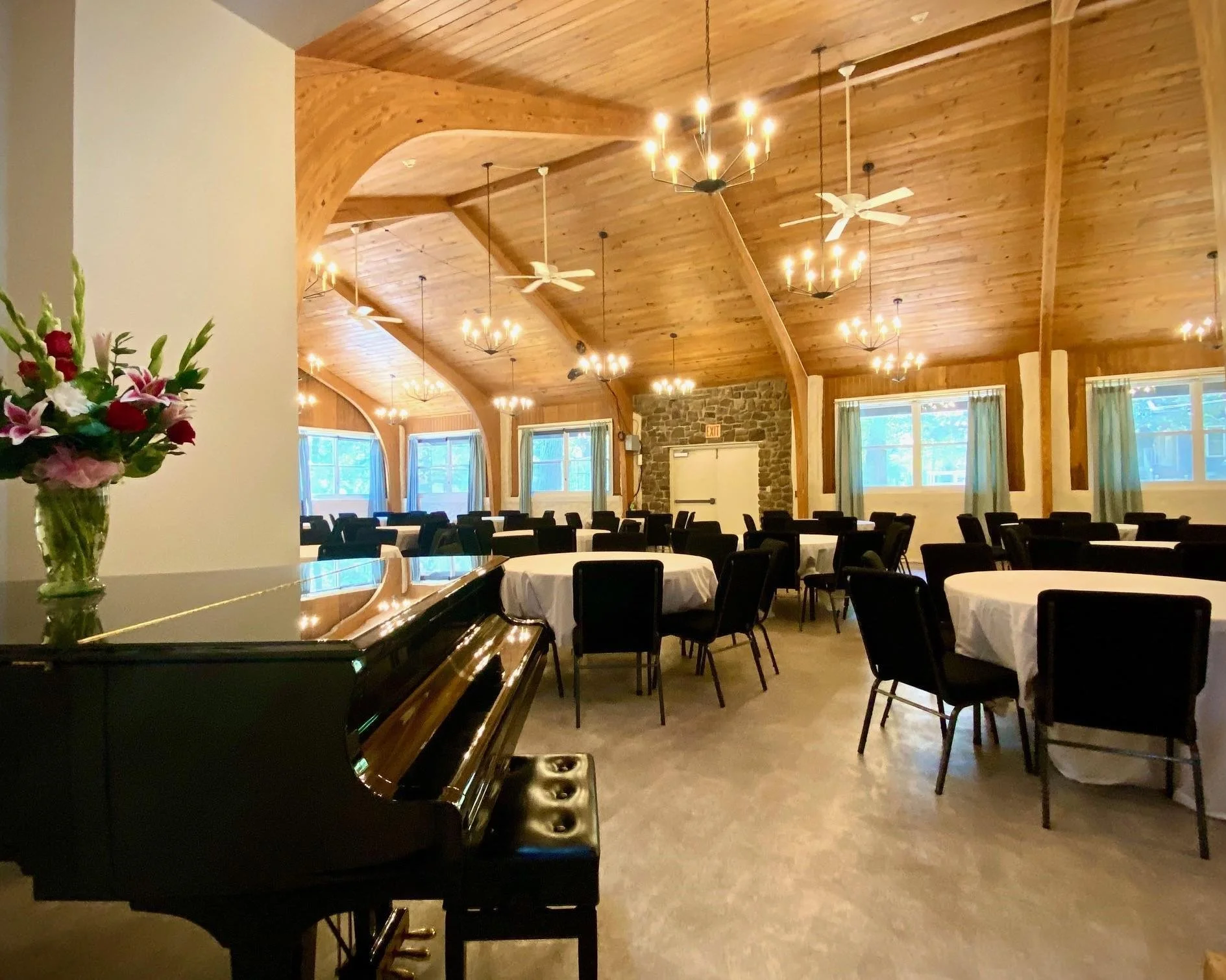 Interior of a wooden banquet hall with round tables, chairs, a stone fireplace, ceiling fans, chandeliers, and windows with curtains, decorated for an event.