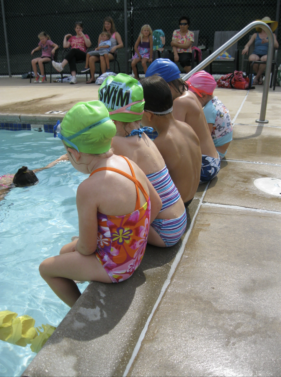 Children sitting on the edge of a swimming pool wearing swimsuits and swim caps, watching a swimming lesson or activity, with spectators seated in the background.