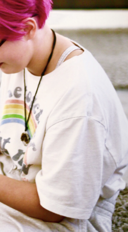 Close-up of a person with short pink hair, wearing a white T-shirt with a rainbow, and a necklace with a pendant.
