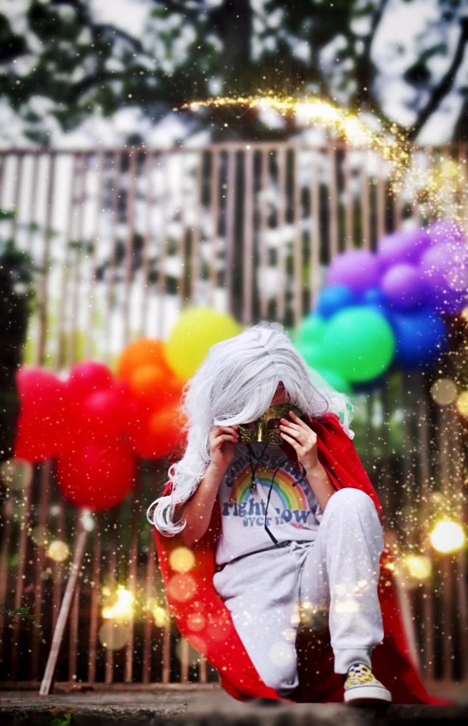 Child dressed in a rainbow t-shirt and white pants, wearing a wig and a cape, sitting on a swing with colorful balloons in the background, holding a camera in front of their face, surrounded by a sparkle effect.