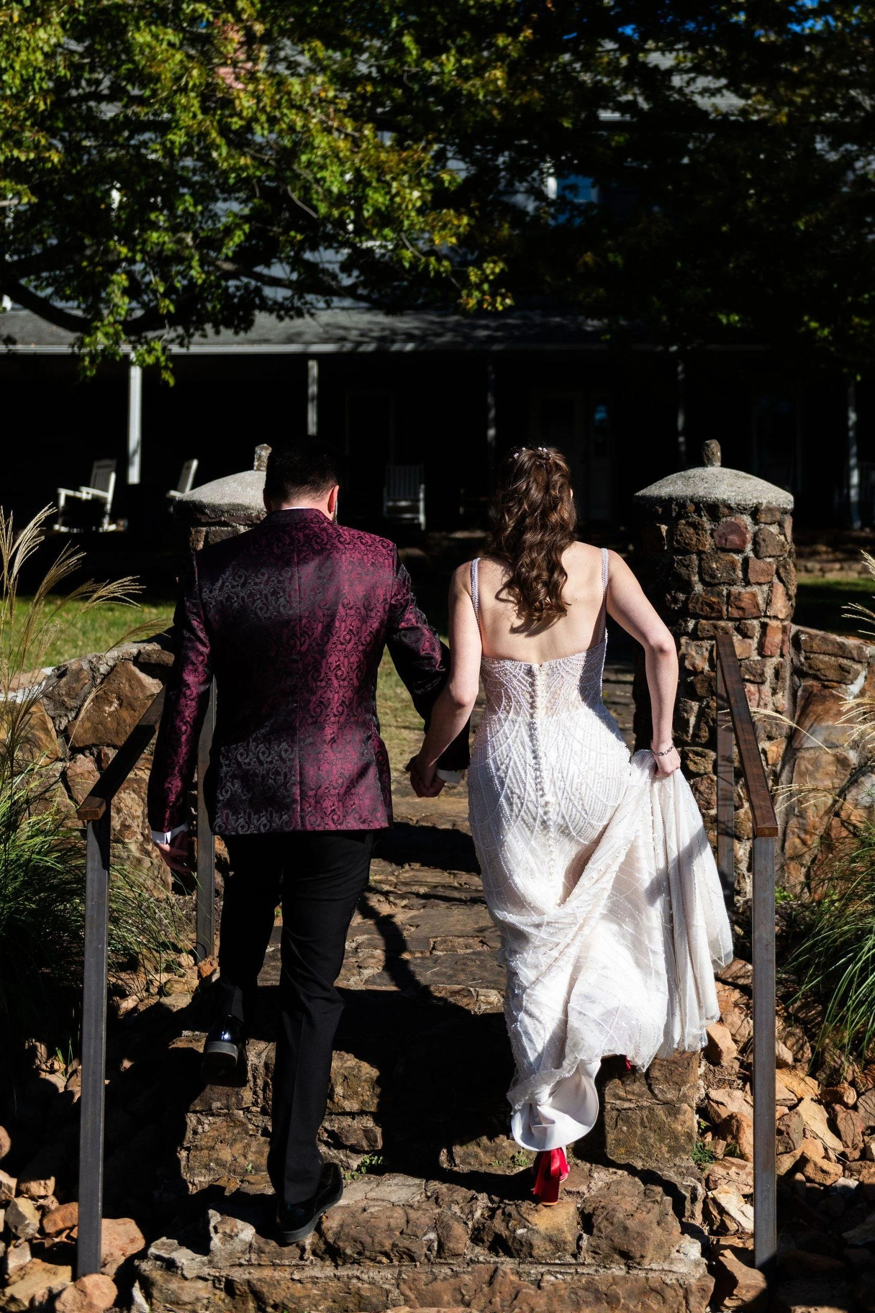 A couple dressed in formal attire, holding hands and walking up stone steps on a sunny day, with trees and a house in the background.