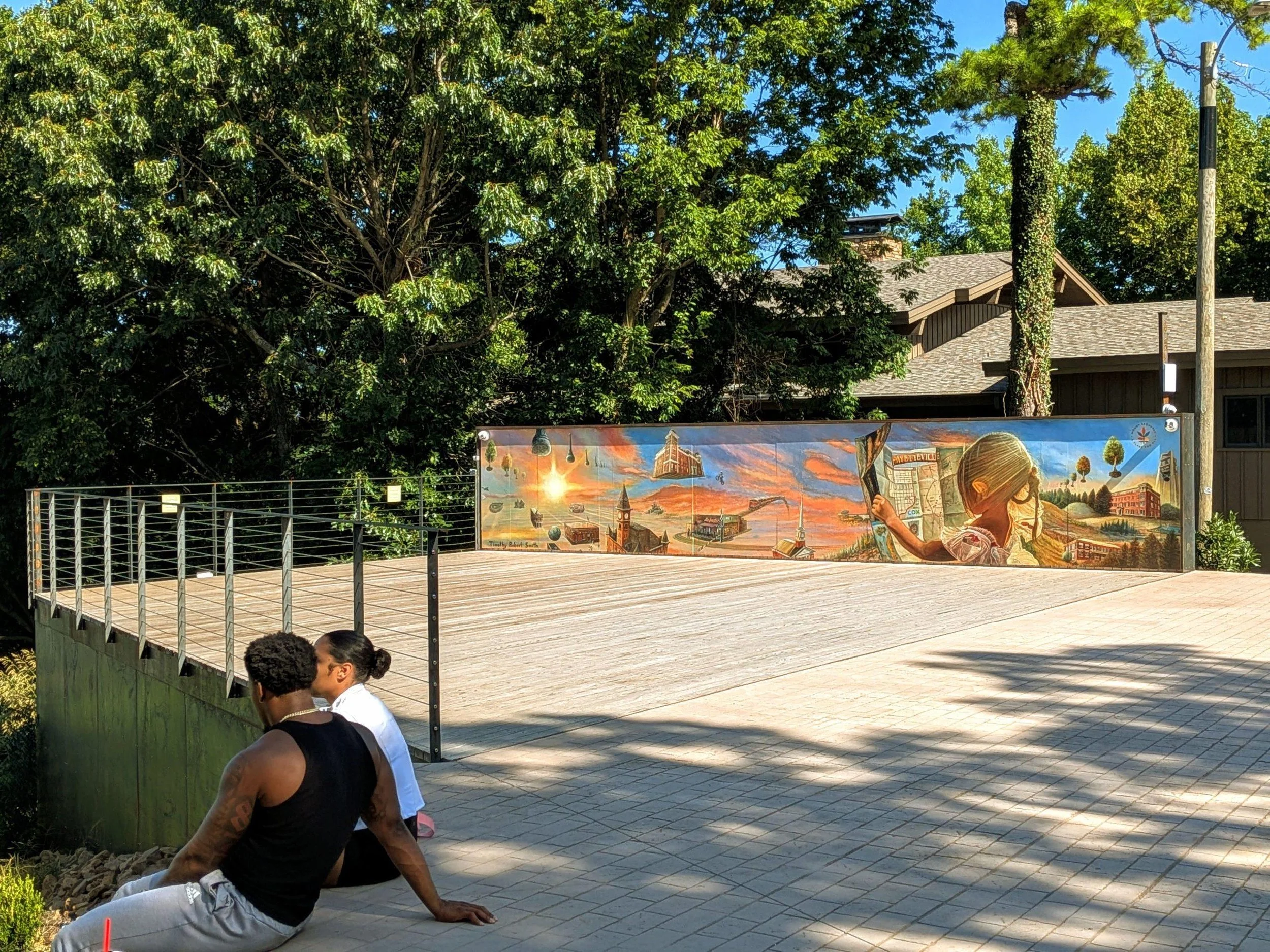 People sitting on outdoor patio in front of a colorful mural with cityscape and bright sunset scene, surrounded by trees and a wooden building.