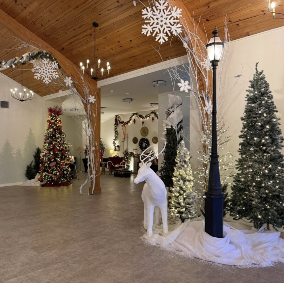 Festively decorated indoor space with Christmas trees, a white reindeer figure, snowflake ornaments, a street lamp prop, and holiday garlands, creating a cozy holiday atmosphere.