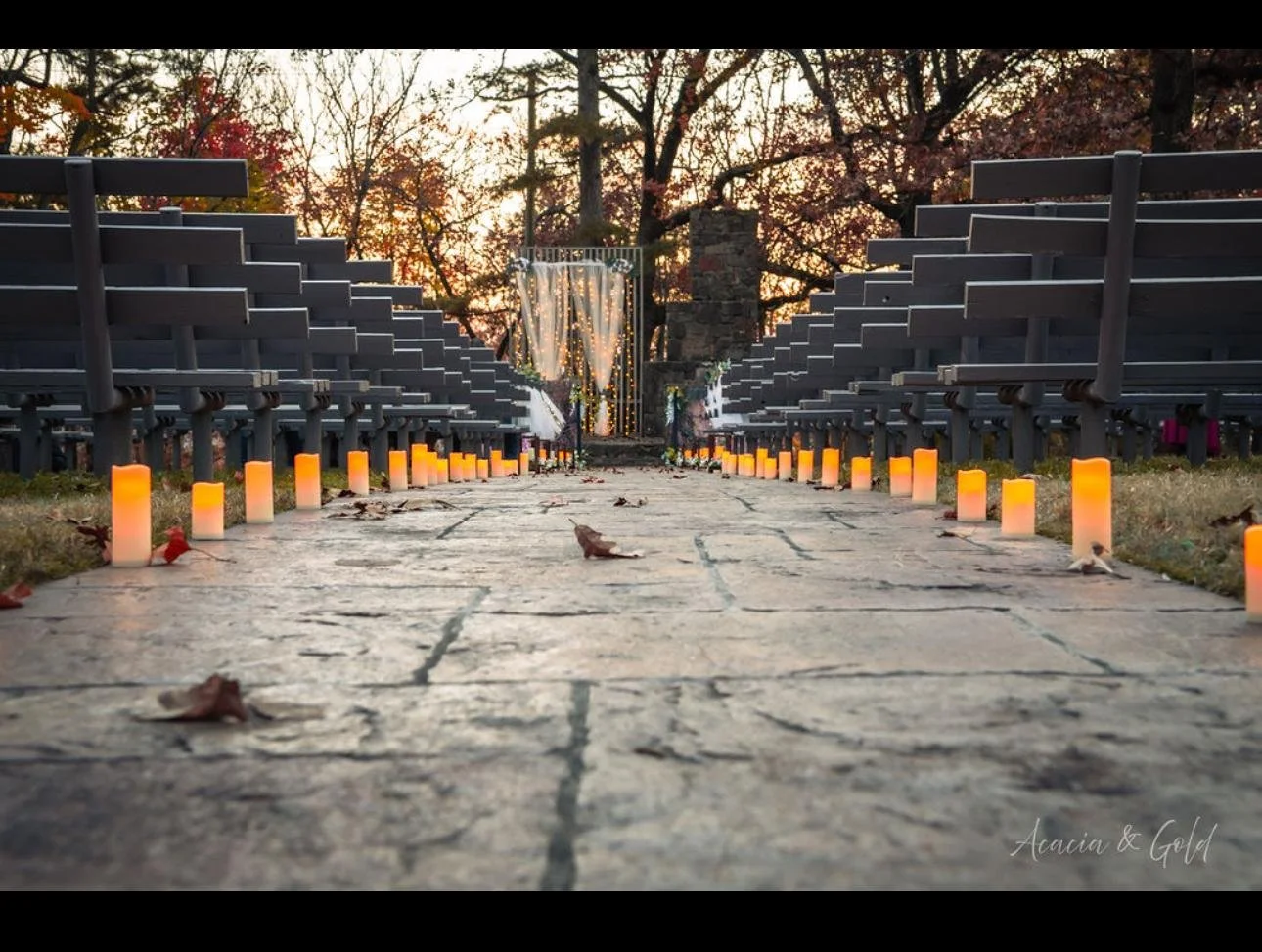 A walkway lined with orange candles leading to an outdoor wedding ceremony setup with chairs on either side, decorated with string lights and drapes at sunset.