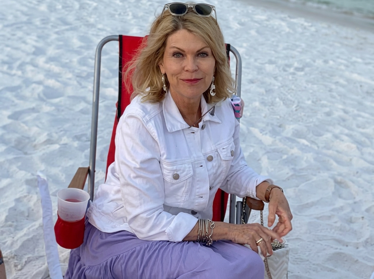 A woman with blonde hair sitting on a red beach chair on the sand, wearing a white jacket, purple skirt, sunglasses on her head, and jewelry, with a drink in a cup holder attached to her chair.