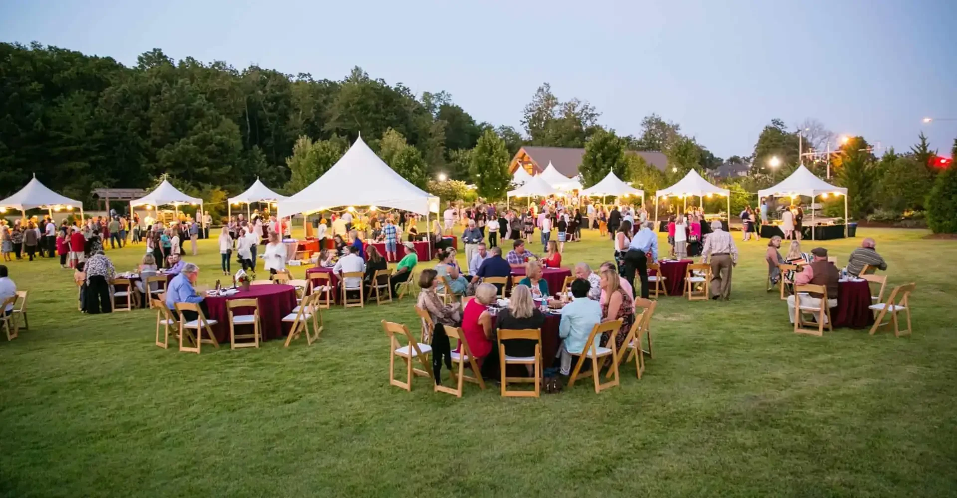 Outdoor event at dusk with numerous people gathered around round tables with maroon tablecloths, seating on wooden chairs on a grassy area. Several white canopies and tents are set up, with food and drink stations. The crowd includes adults socializing and walking around, with trees and a building in the background. The scene is illuminated by string lights and outdoor lighting.