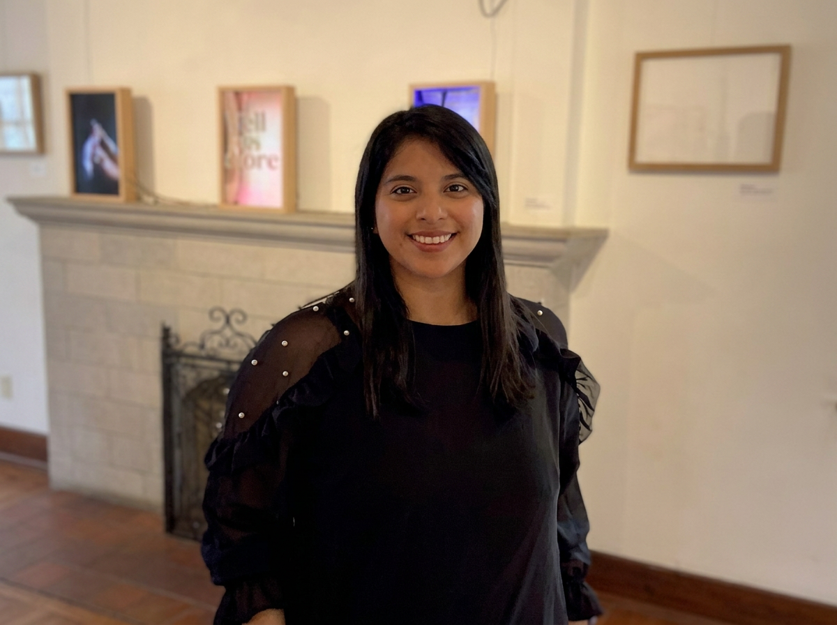 A woman with shoulder-length dark hair and a black top with pearl embellishments on the shoulders, smiling indoors in front of a fireplace with artwork on the wall.