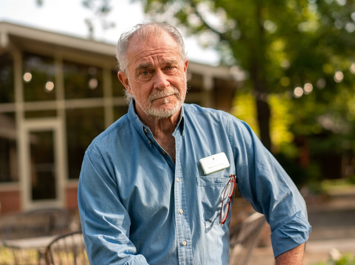 An elderly man with gray hair and a beard wearing a blue button-up shirt, standing outdoors near a building with trees in the background.
