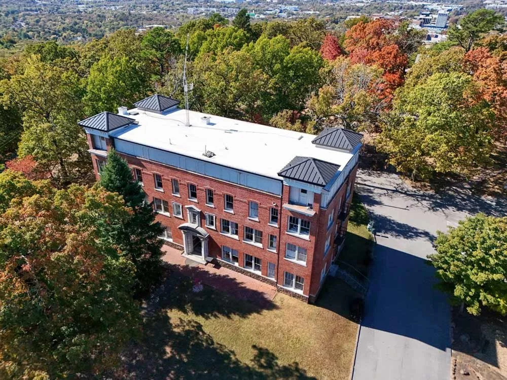 Aerial view of a multi-story red brick building surrounded by trees with fall foliage. The building has a white roof and black metal accents on the roof edges. Trees and a parking lot are nearby.