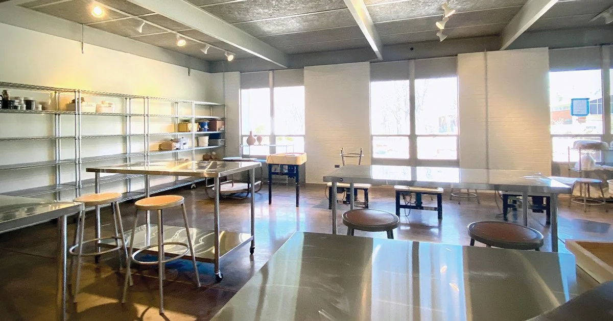 Empty commercial kitchen or cafe with stainless steel tables, shelves with dishes and bowls, stools, and large windows letting in natural light.