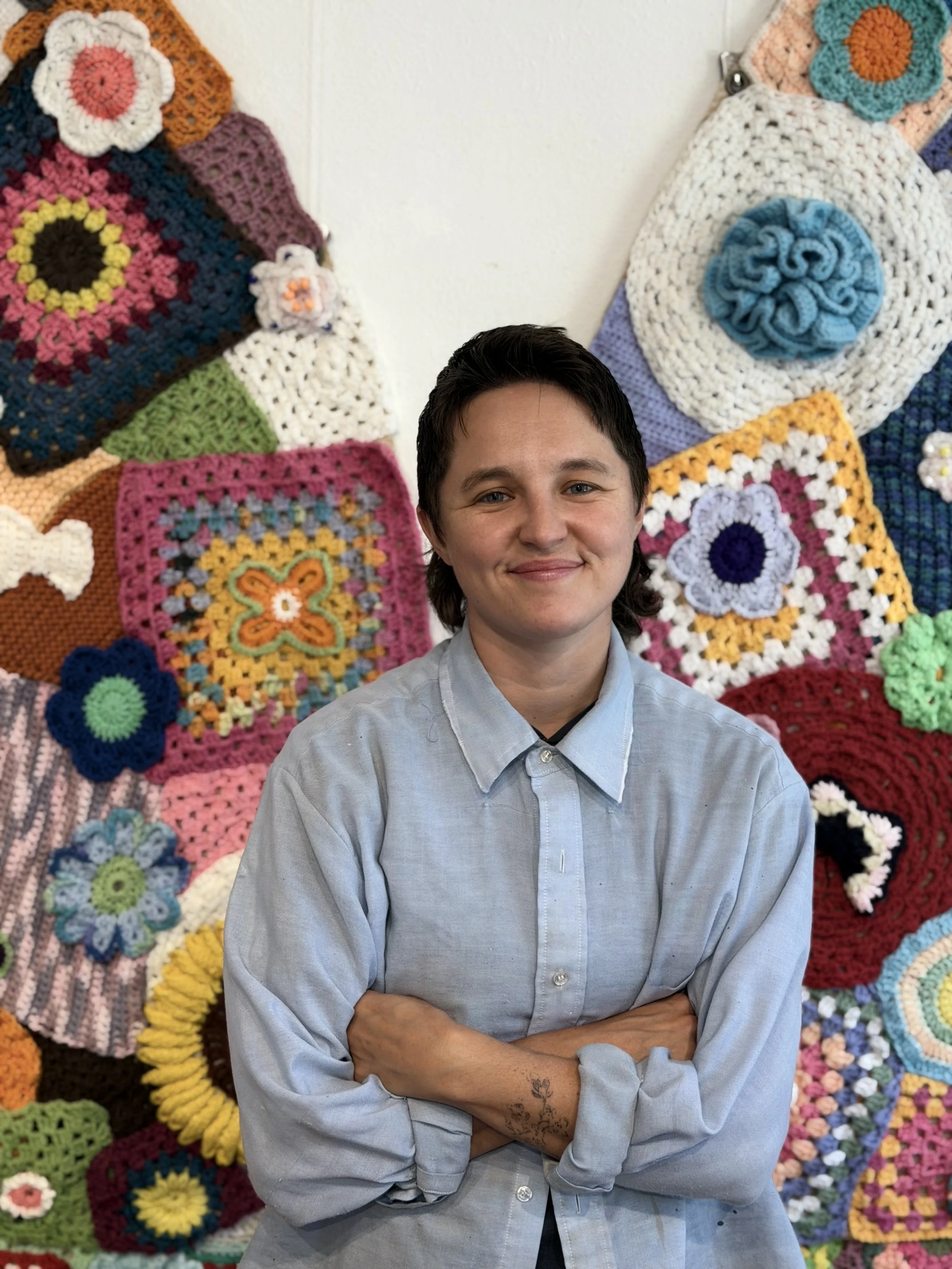 A young person with short dark hair and tattoos on their arm standing in front of a colorful wall of crocheted blankets and granny squares.