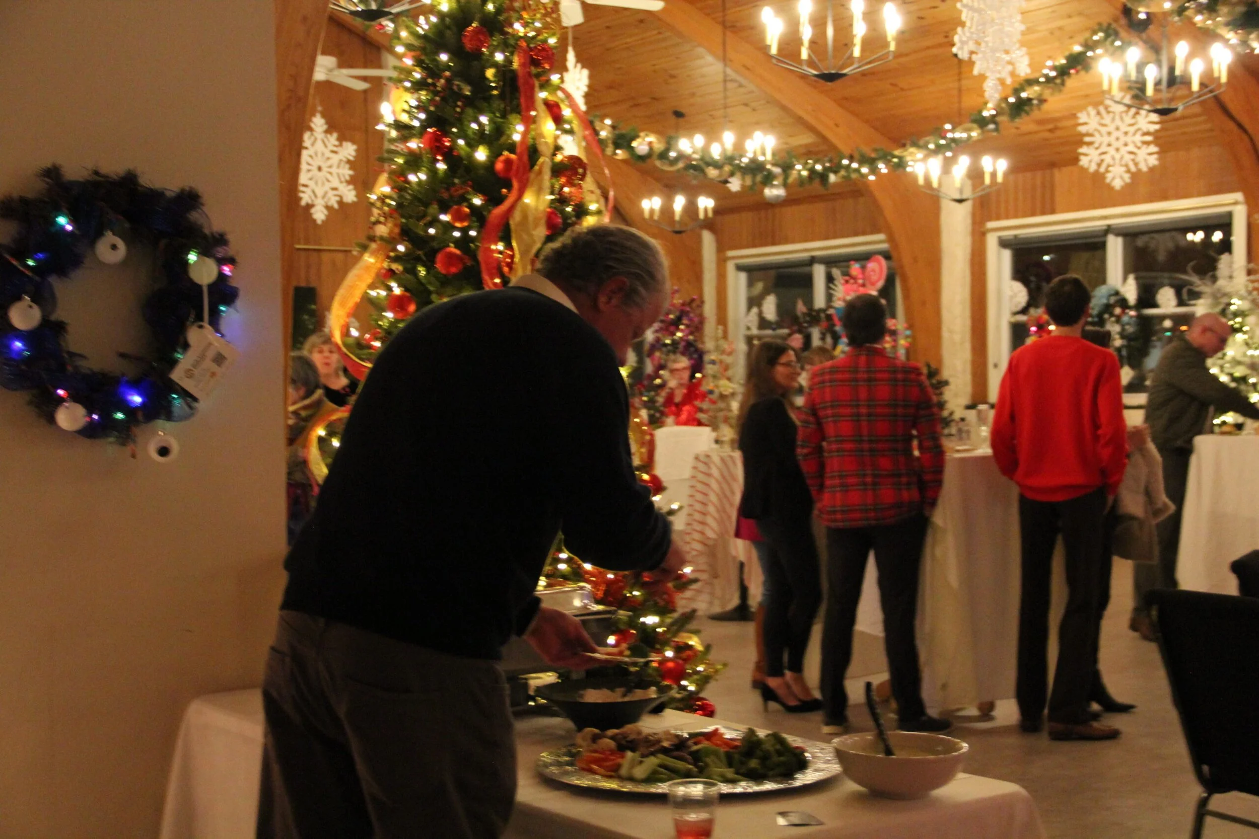 A Christmas gathering with a decorated tree, people mingling, and holiday decorations in a cozy, wood-paneled room.