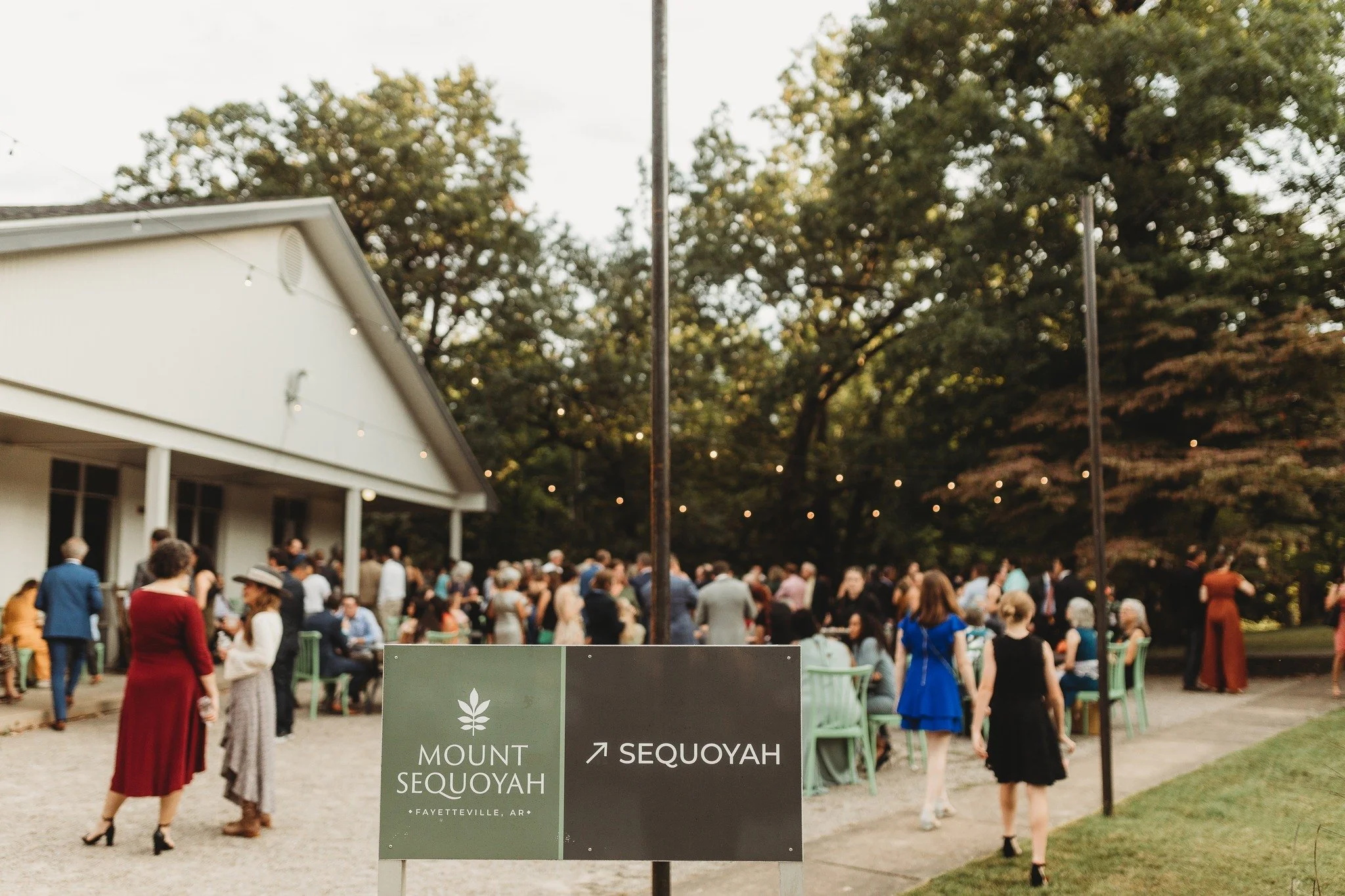 Outdoor wedding reception with many guests gathered near a white building, outdoors with green trees in the background. There are string lights hanging overhead, and a sign with directions to Mount Sequoyah and Sequoyah in Fayetteville, Arkansas.