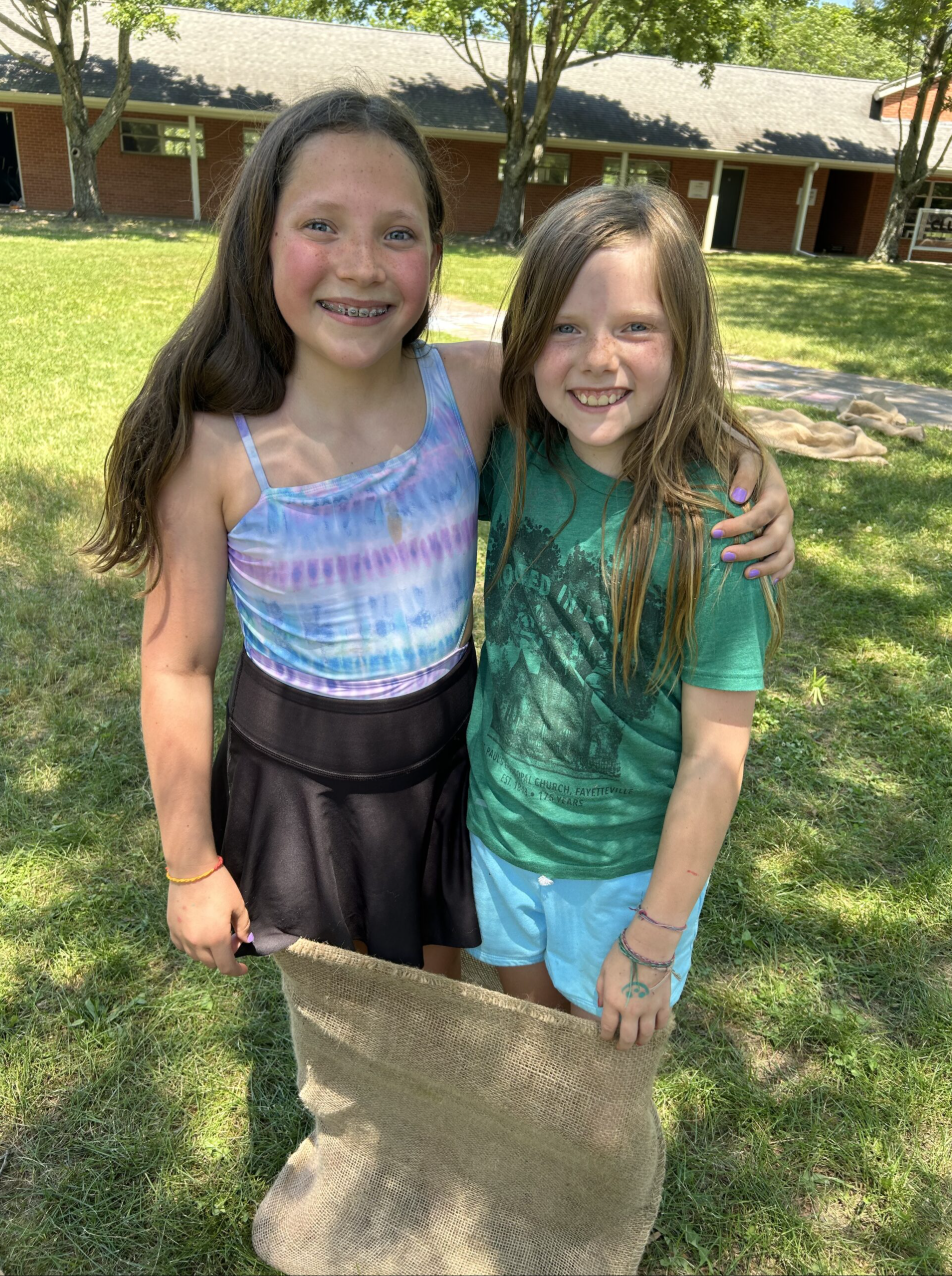 Two young girls smiling and embracing each other outdoors on a sunny day, with trees and a building in the background, holding a large burlap sack.