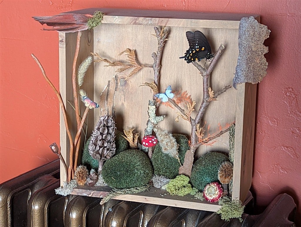 A handcrafted nature scene with artificial trees, moss, and mushrooms in a wooden frame, featuring butterflies and moss decorating the display.