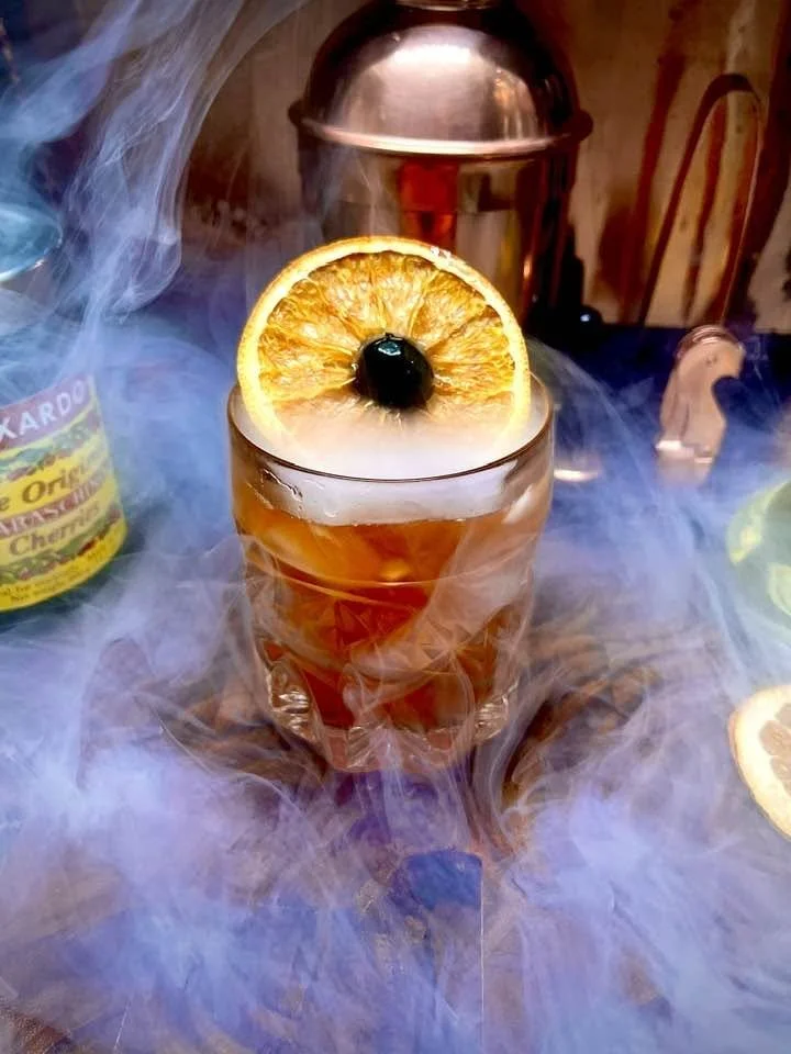 A glass of cocktail garnished with a slice of orange and a black cherry, surrounded by smoke, on a wooden table with a vintage bar setting in the background.