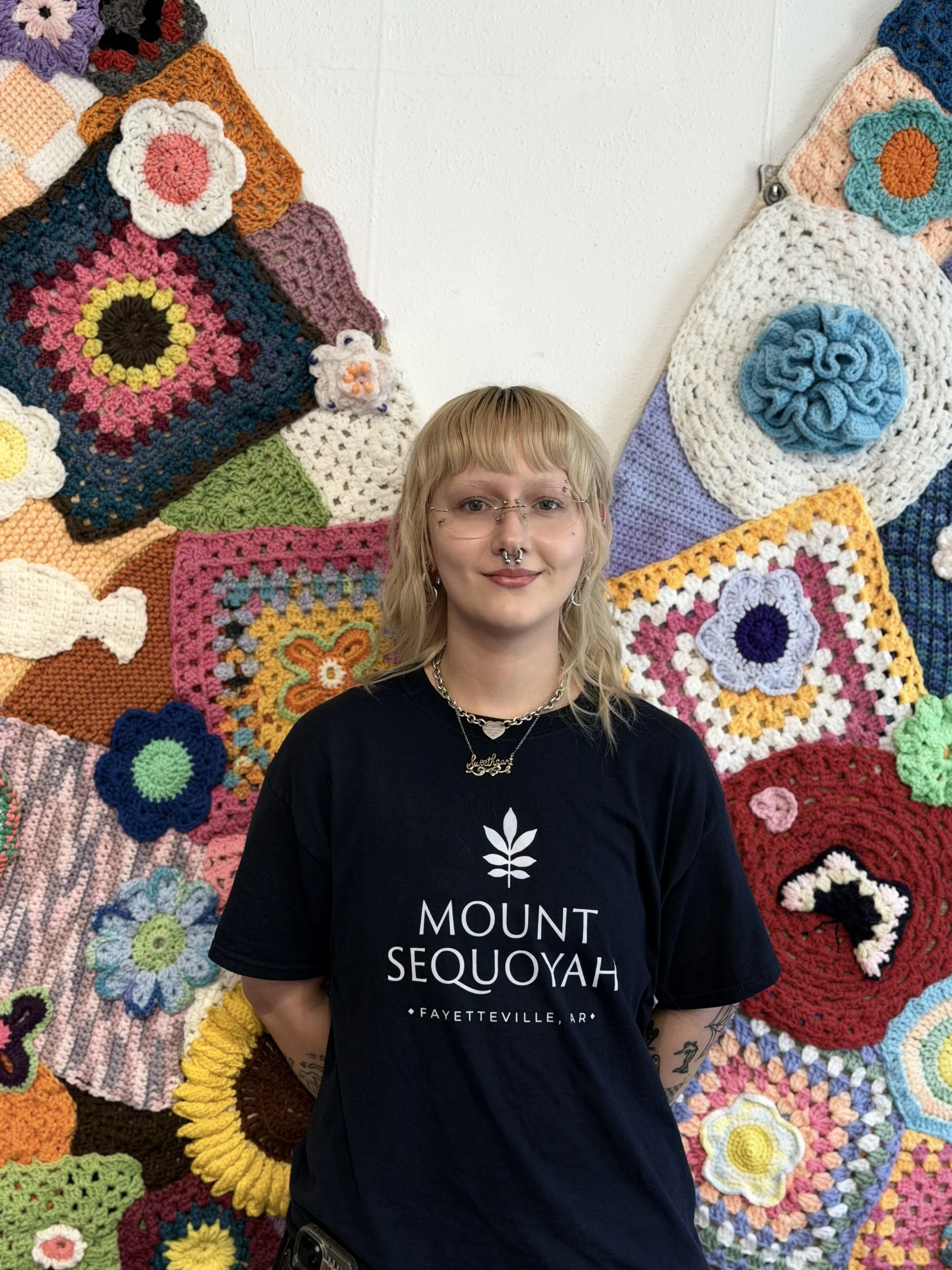 A person with blonde hair and glasses standing in front of a wall decorated with multicolored crochet blankets. They are wearing a black t-shirt with white text reading 'MOUNT SEQUOYAH FAYETTEVILLE, AR'.