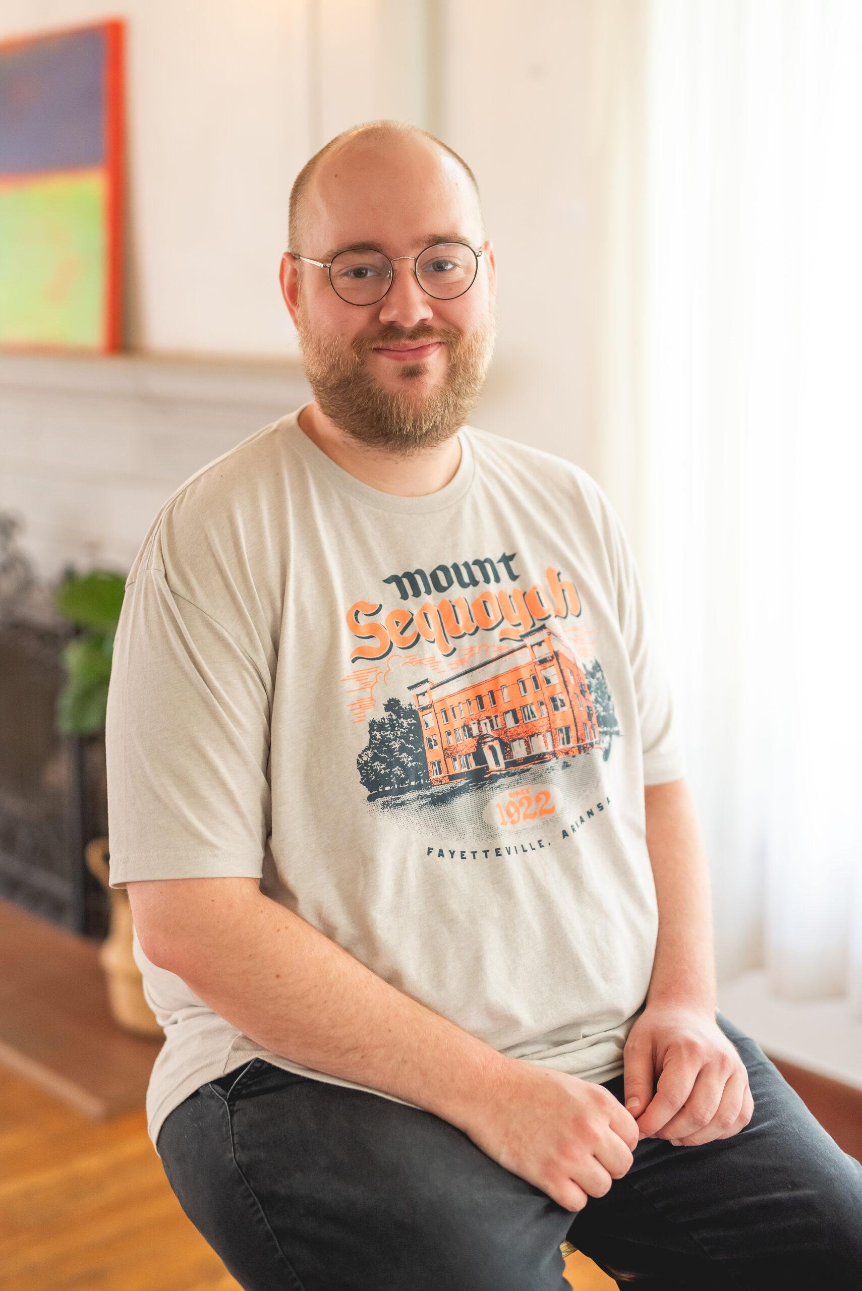 A young man with glasses and a beard is sitting on a chair indoors. He is wearing a beige T-shirt with a graphic of a building and text that reads 'Mount Sequan.' He has a slight smile and is looking at the camera. The background shows a bright room with white walls and some colorful items on the wall.
