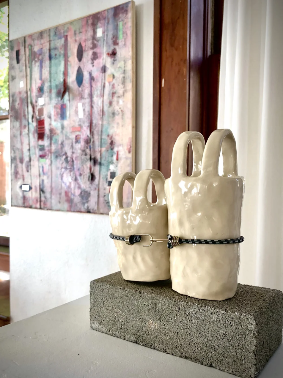Two ceramic ceramic vases shaped like bunny heads with handles, connected by a black cord, placed on a gray stone block. In the background, a colorful abstract painting hangs on the wall in a room with a window and wooden furnishings.