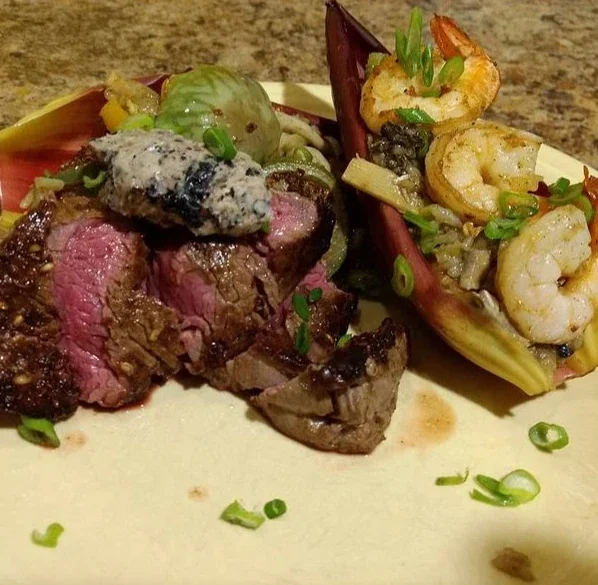 Cooked steak cooked to medium rare, topped with a blue cheese sauce, served with a side salad with mixed greens and a shrimp taco filled with shrimp, lettuce, and chopped green onions.