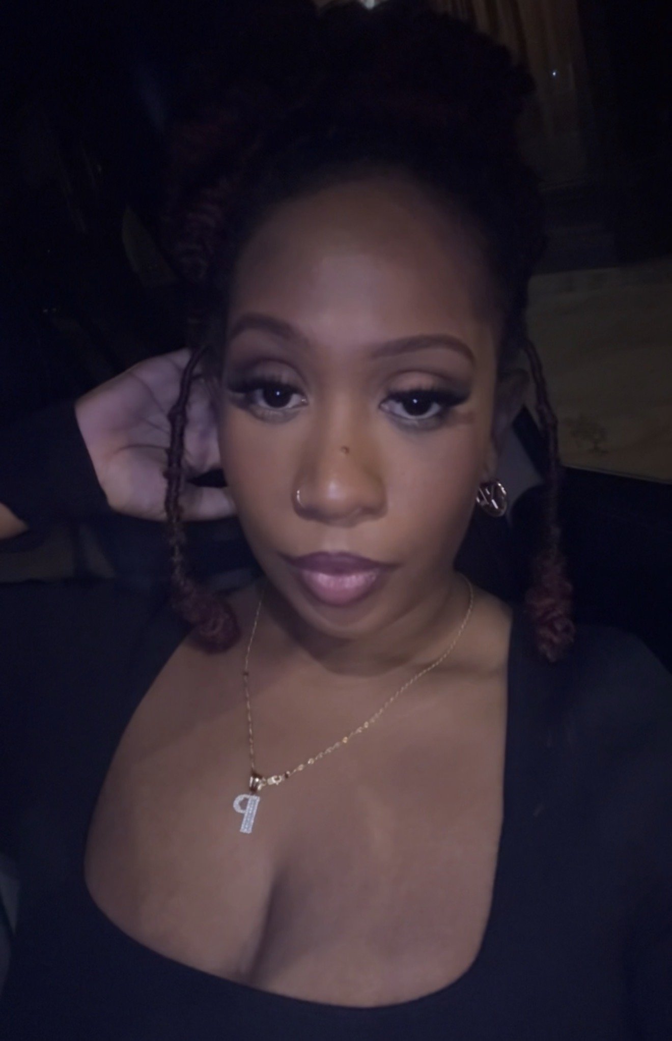 A close-up of a woman with makeup, wearing a black top with a deep neckline, a gold necklace with a letter 'P' pendant, and earrings. She has her hand behind her head and is looking into the camera.