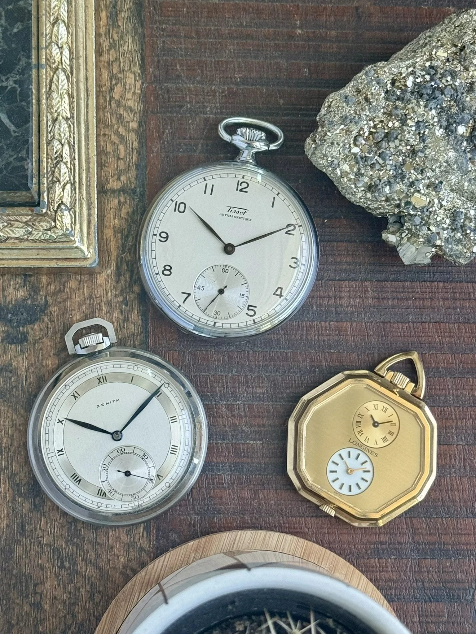 3 vintage pocket watches sitting on a table: a Longines dual time pocket watch in a gold-tone case, a Tissot, and a Zenith sector dial.
