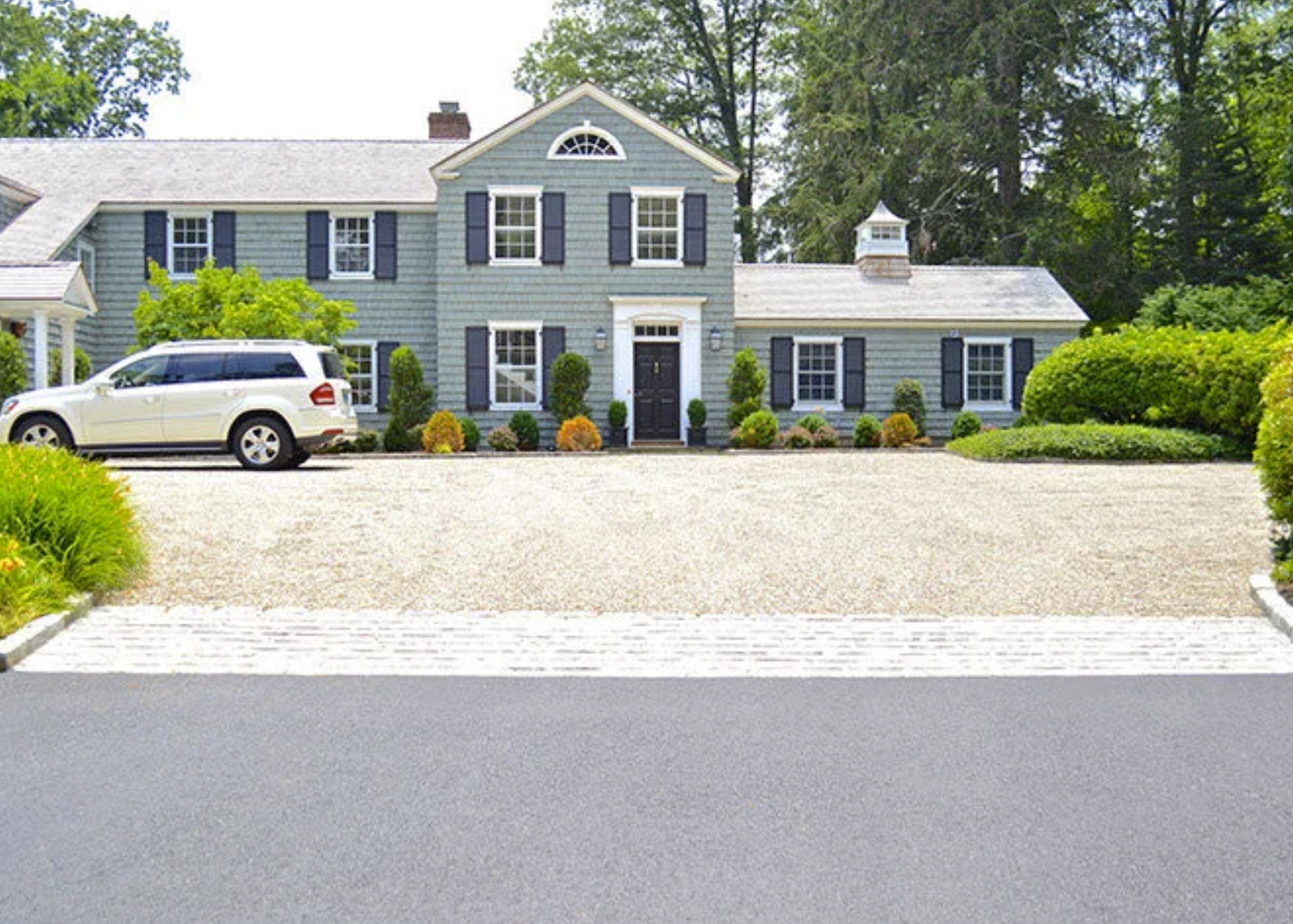 Front view of a two-story house with blue siding, black shutters, and a black front door, surrounded by green trees and landscaped bushes, and a white SUV parked in the driveway.
