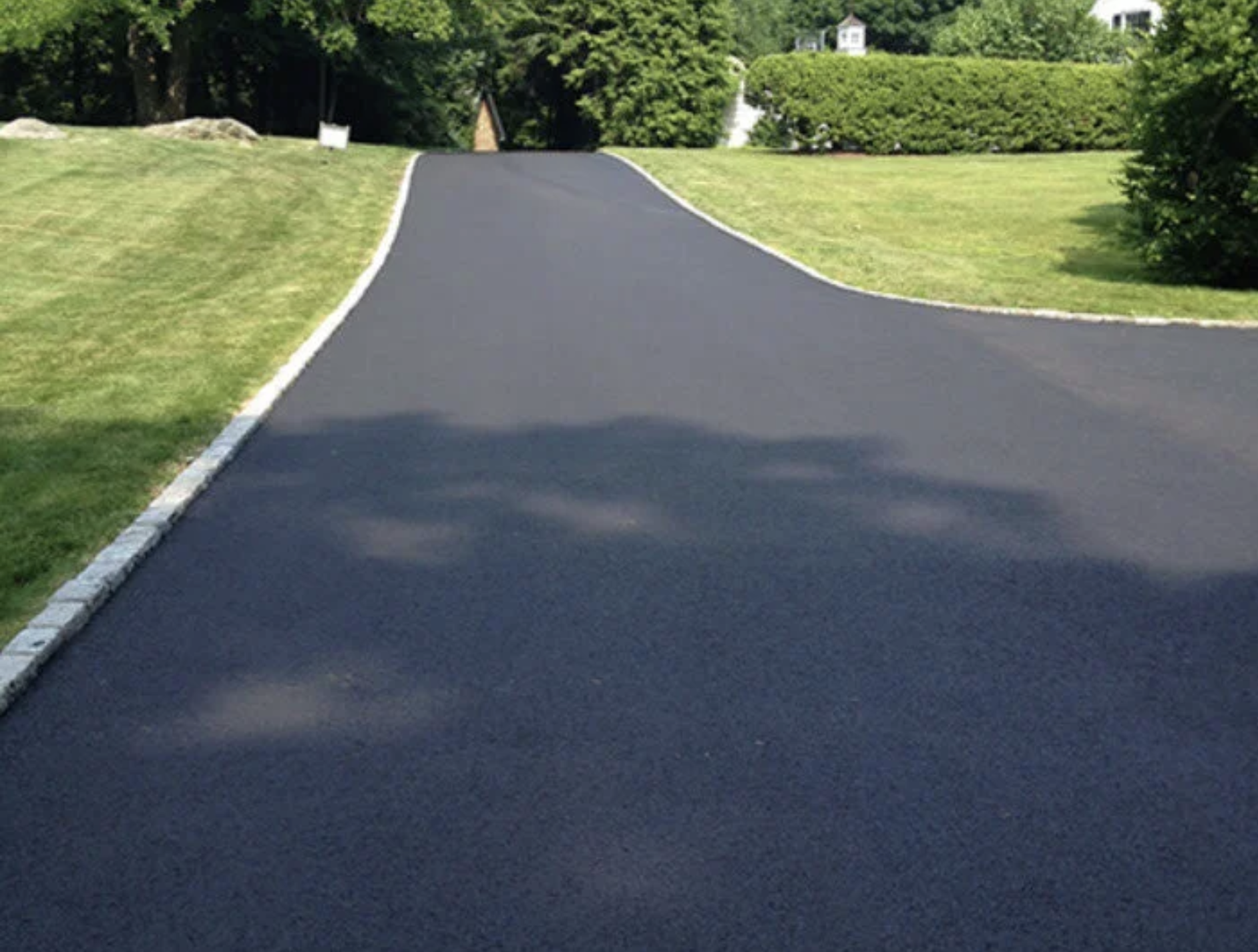 A paved driveway with a white border, surrounded by well-kept green grass and lush trees, leading to a residential area in the background.
