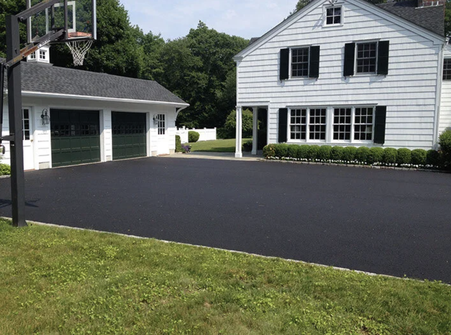 A residential backyard with a basketball hoop, a large white house with black shutters, a green lawn, and a freshly paved driveway.