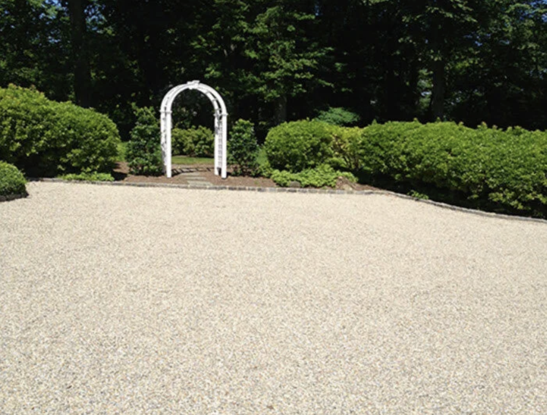 A garden with a white decorative archway, green bushes, and trees in the background.