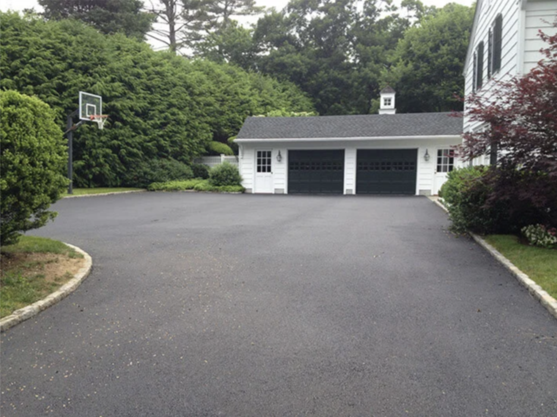 A paved driveway in a residential area with a basketball hoop on the left and a white house with black garage doors on the right, surrounded by green trees and bushes.