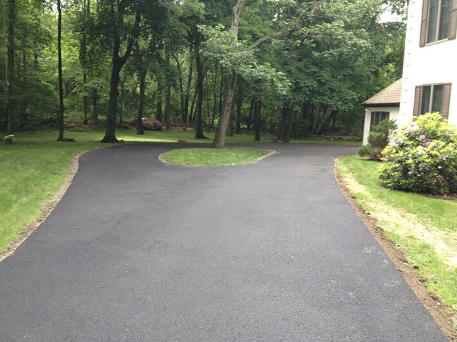 A freshly paved asphalt driveway in front of a house, bordered by grass and lined with bricks. The background features a wooded area with tall trees and dense green foliage.
