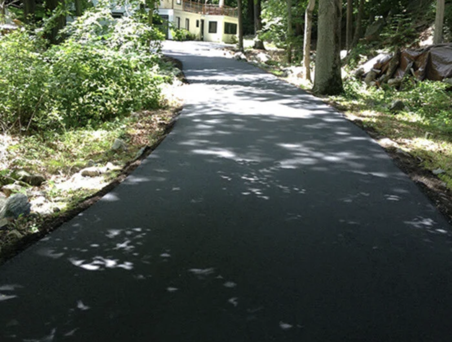 A paved sidewalk winding through a wooded residential area with a house in the background, surrounded by trees and greenery, with shadows cast on the path.