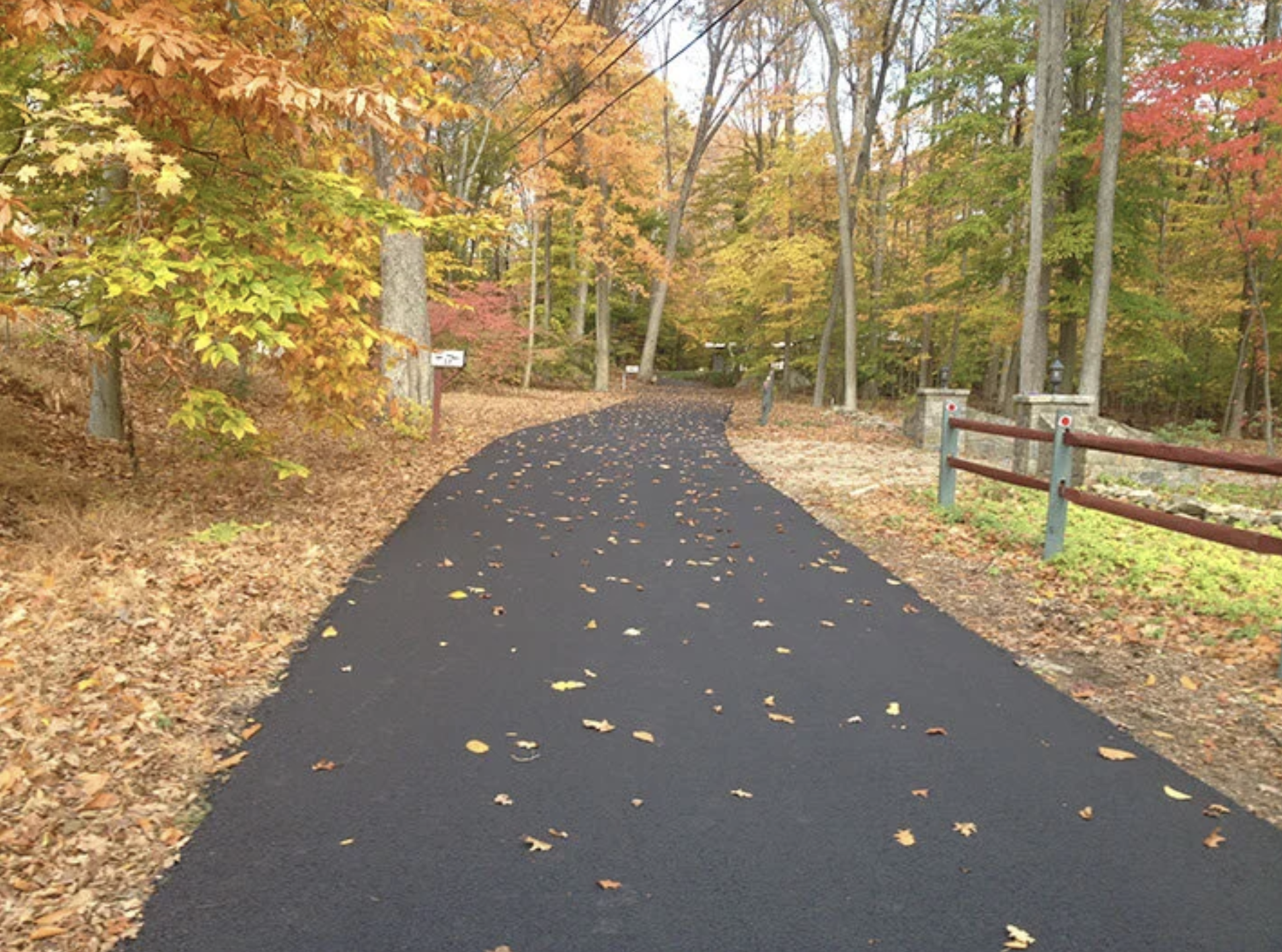 A paved blacktop path winding through a wooded park during fall, surrounded by colorful autumn leaves on trees and on the ground, with utility wires overhead and stone posts with a red and white fence on the right side.