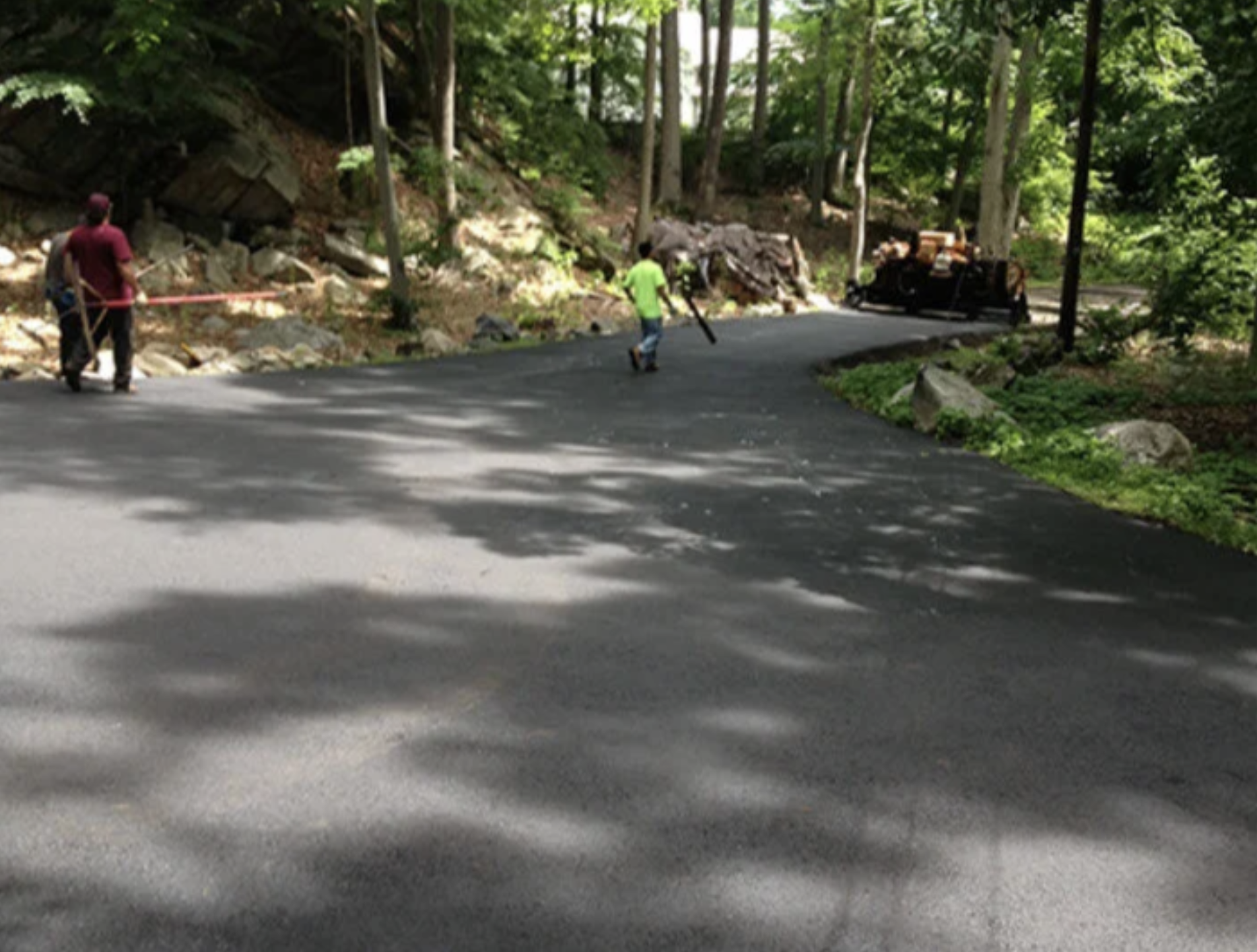 A residential street with trees and a hillside. Two people are standing by the curb, one wearing a maroon shirt and the other a bright green shirt. A black vehicle appears to be damaged and is parked near rocks and debris on the side of the street.